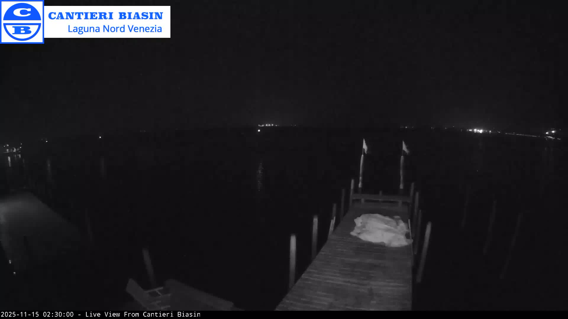 A dark, clear night scene shows a wooden dock extending into calm water, with a white tarp or cover on the dock and distant lights twinkling across the horizon.