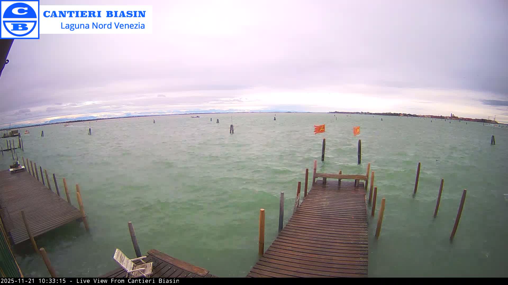 On an overcast day, two wooden docks extend into a choppy, greenish-grey lagoon dotted with numerous channel markers and two orange flags, with a distant shoreline and mountains visible under the cloudy sky.