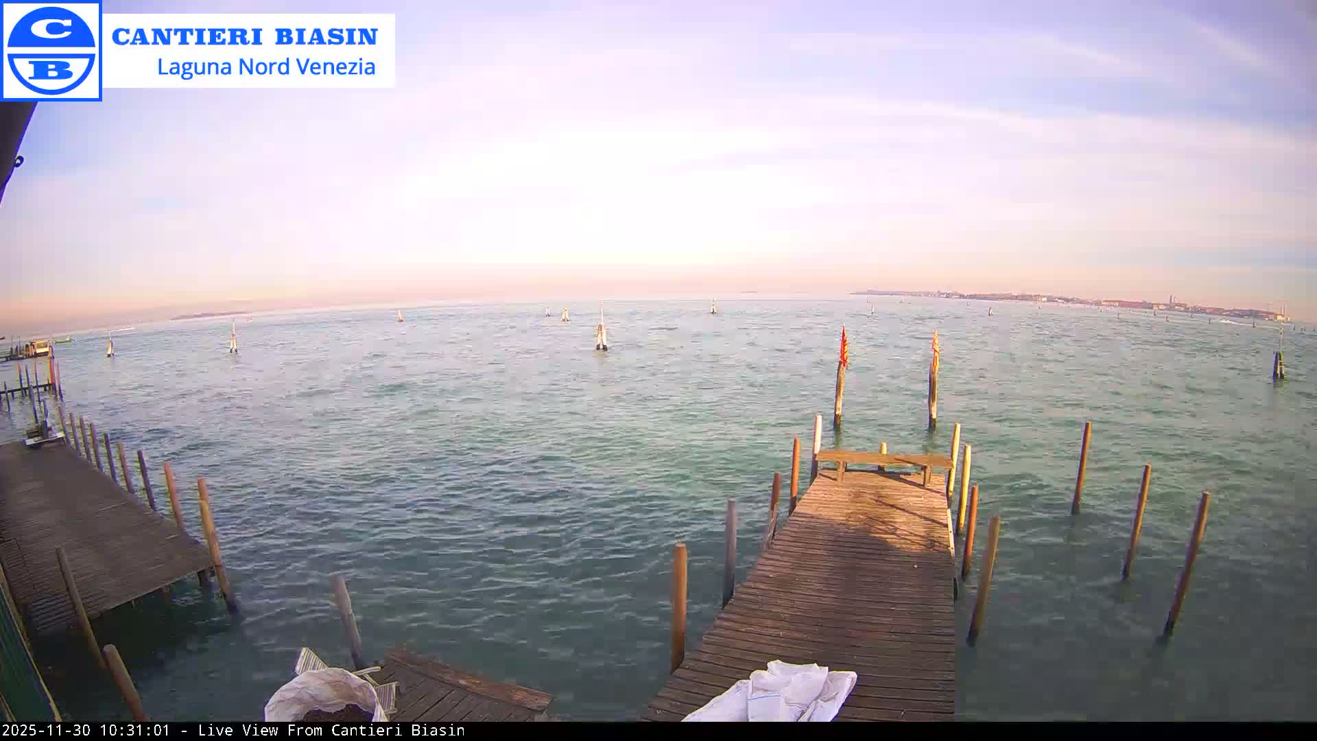 On a clear, bright day, two wooden piers extend into a calm, turquoise lagoon dotted with numerous navigation poles, leading the eye towards a hazy cityscape on the distant horizon.