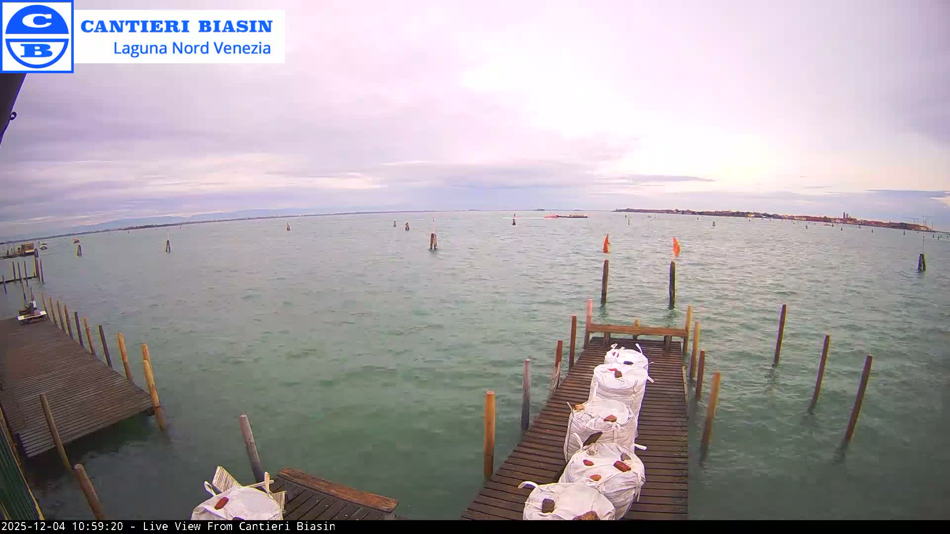 A wide view of a lagoon on an overcast day shows wooden docks laden with large white bags and numerous marker poles in the greenish-blue water, with a distant cityscape visible on the horizon.