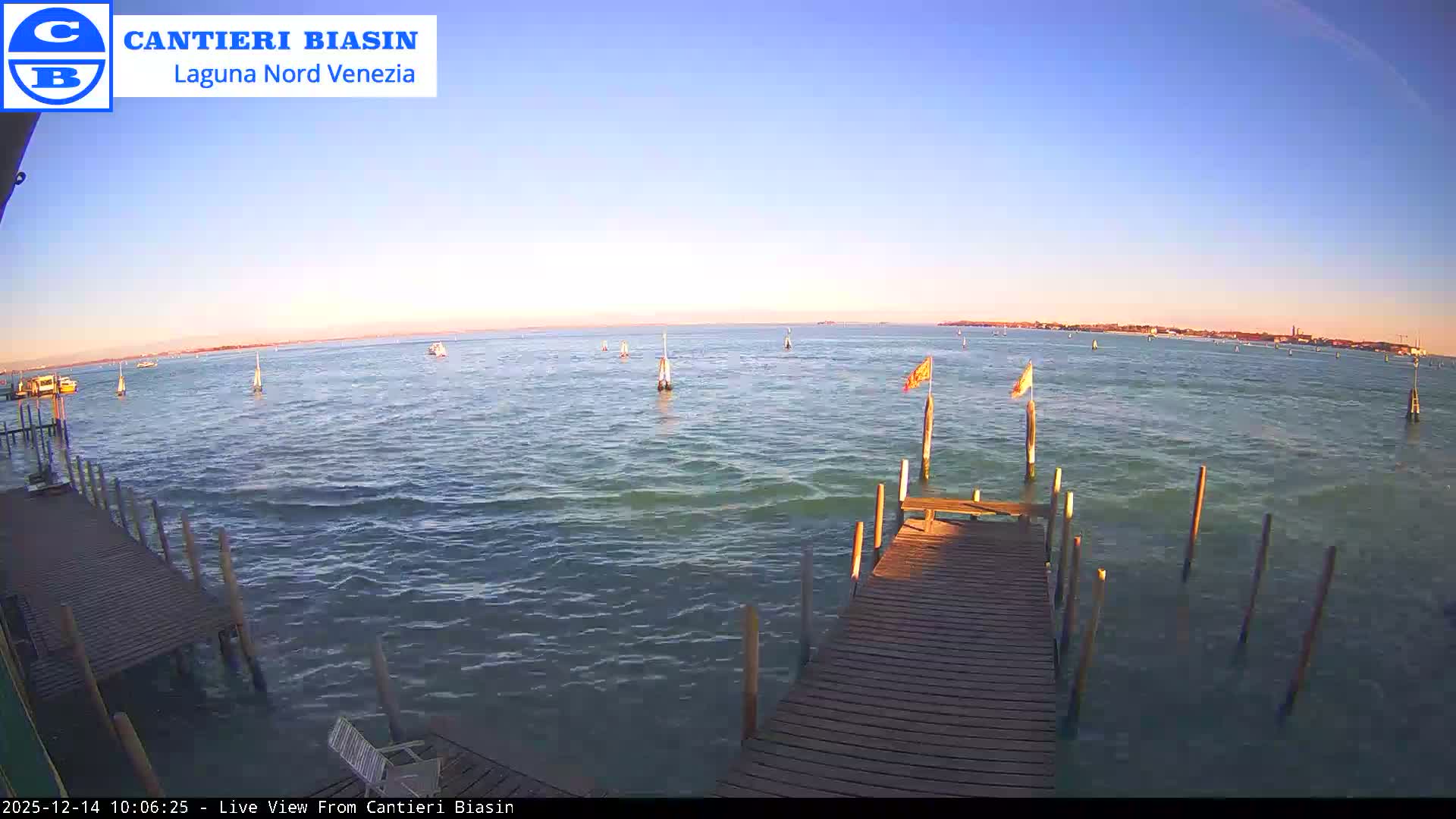 A wide view of a lagoon on an overcast day shows wooden docks laden with large white bags and numerous marker poles in the greenish-blue water, with a distant cityscape visible on the horizon.