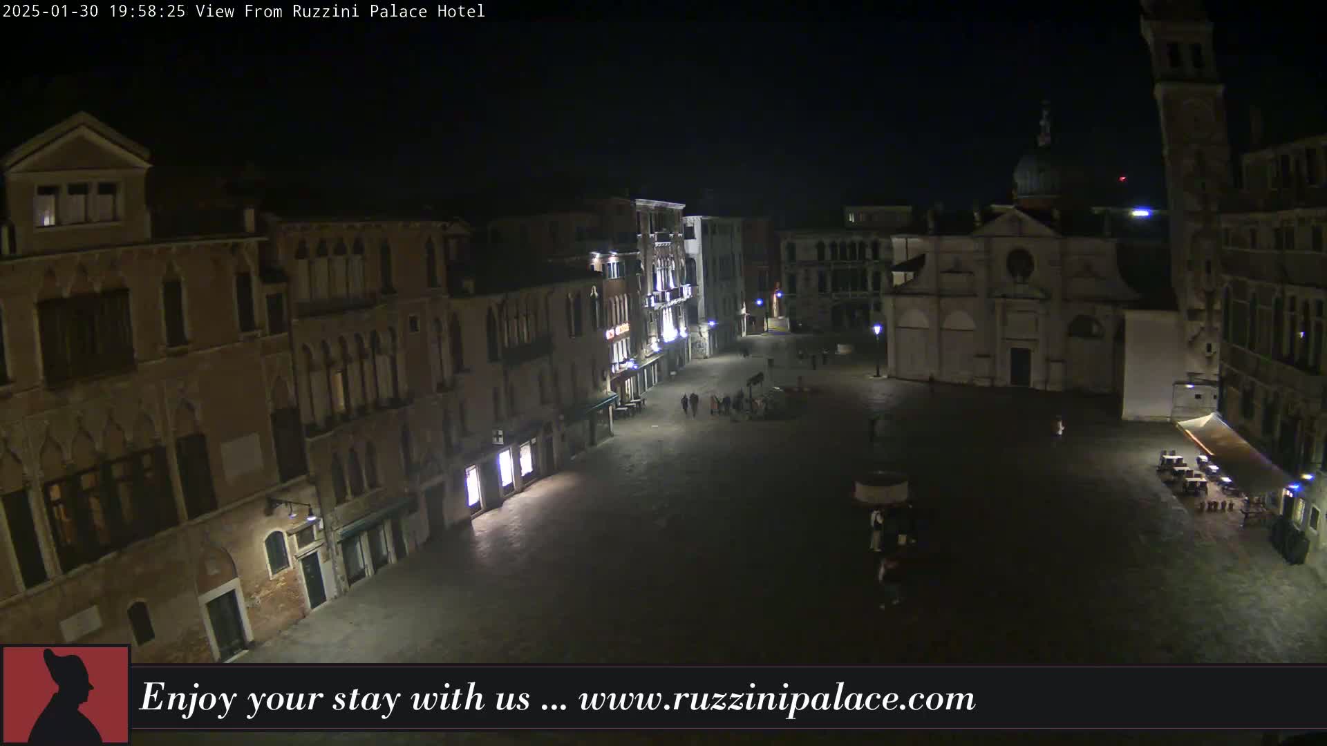 A nighttime, high-angle view of a sparsely populated, illuminated square in Venice, surrounded by historic buildings.