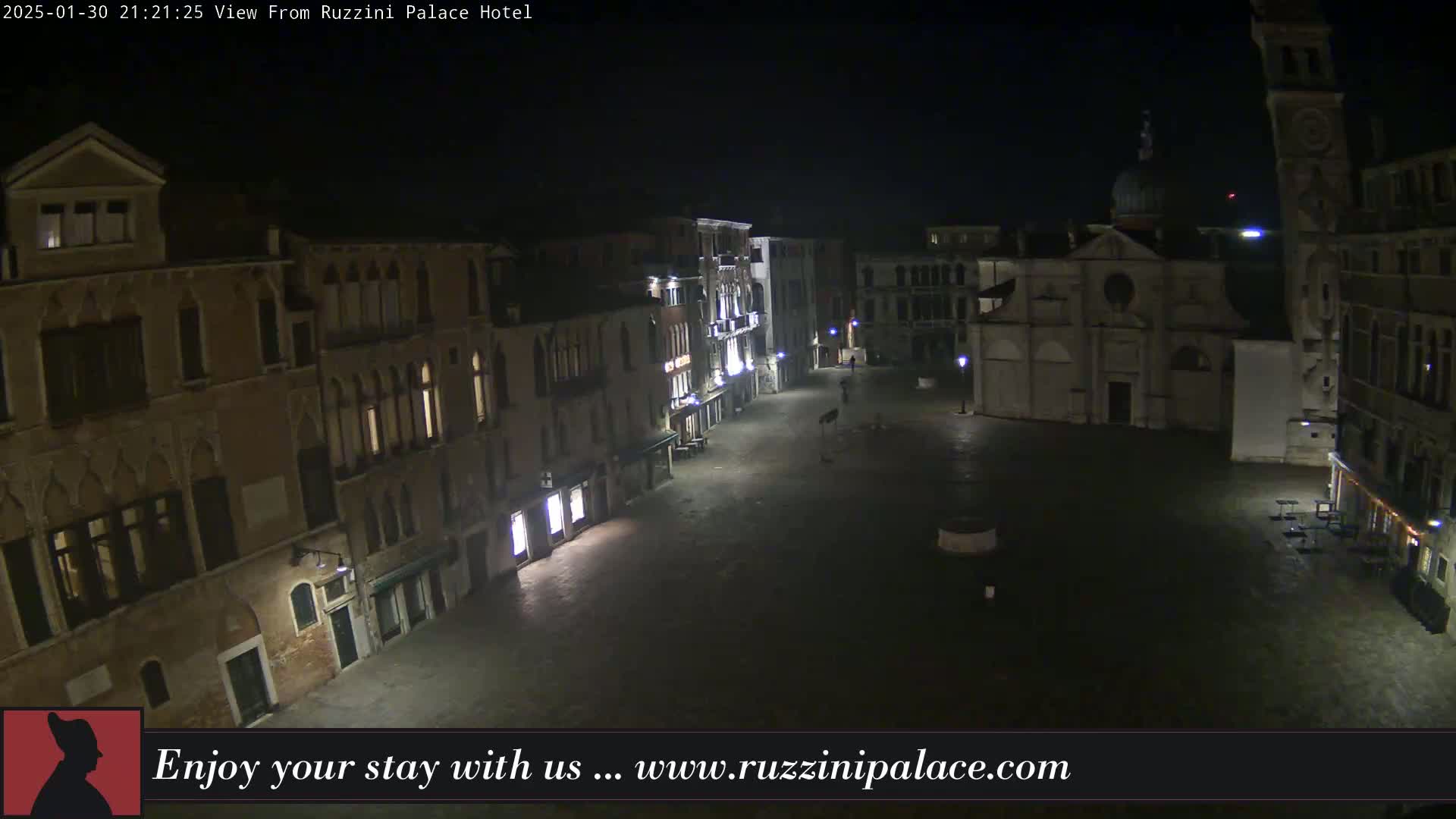 A nighttime, aerial view of a mostly empty, stone plaza in Venice, Italy, with several buildings surrounding it.