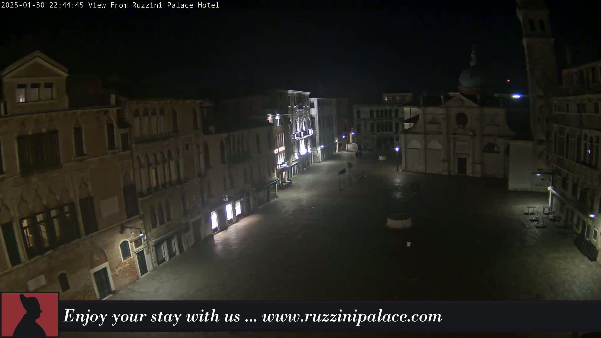 A nighttime view of a mostly empty, cobblestone square in Venice, Italy, surrounded by old buildings and dimly lit by street lamps.