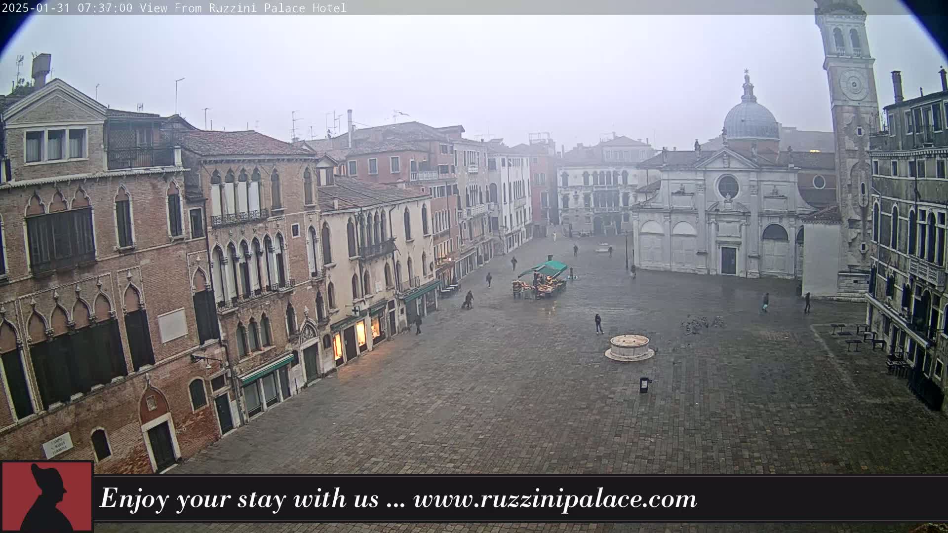 A mostly empty cobblestone square in a foggy city, surrounded by tall, aged buildings, one of which is a church with a dome.