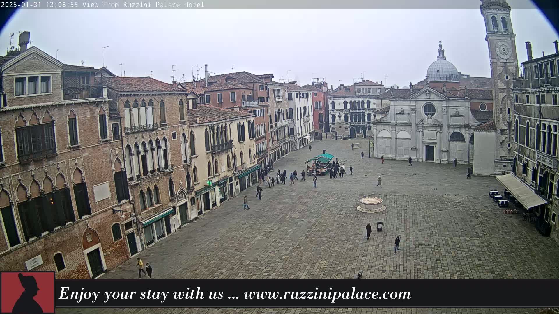 A large, paved square in a city is sparsely populated with people, surrounded by several-story brick buildings under an overcast sky.
