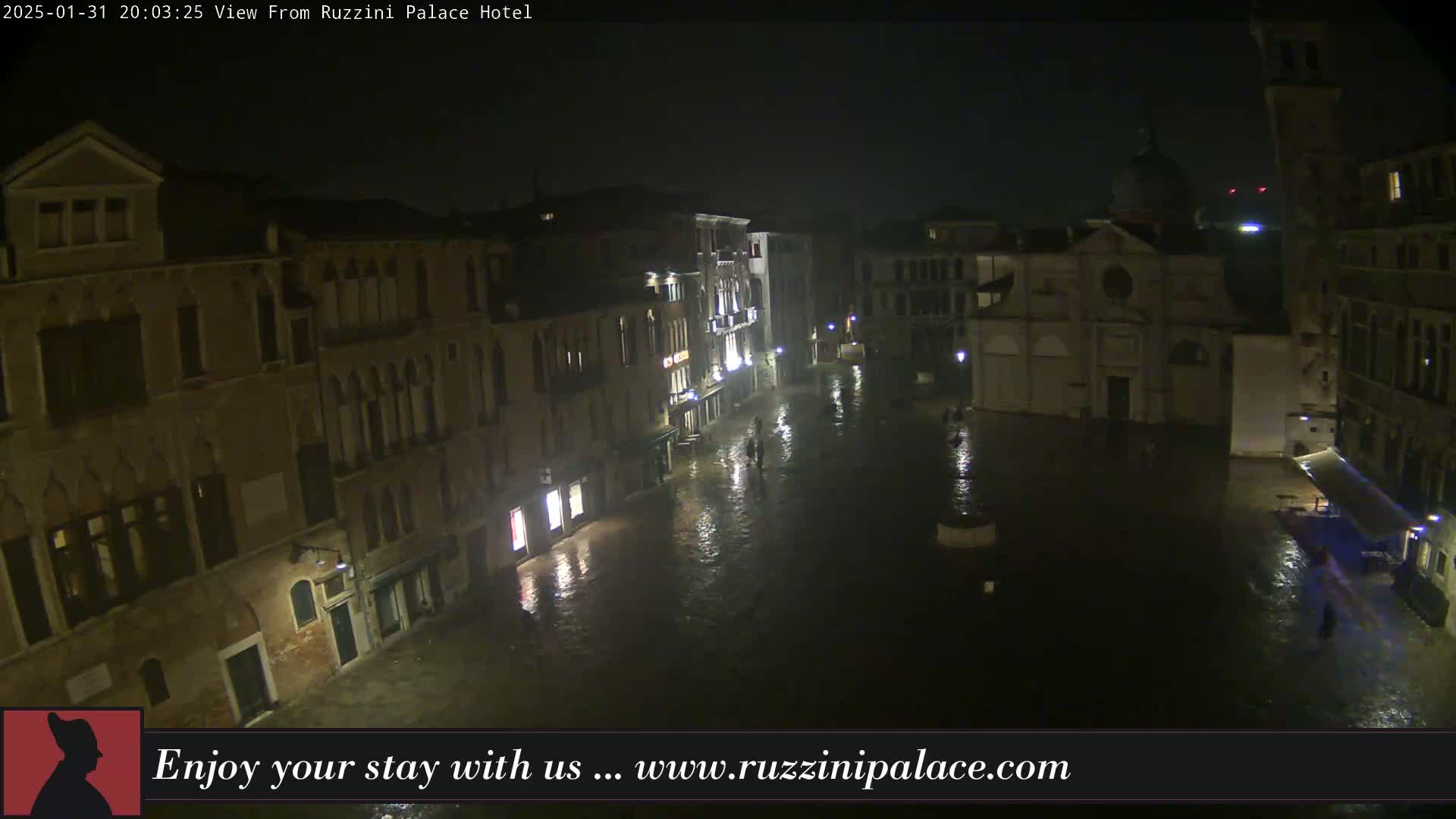 A nighttime view of a rain-soaked Venetian square, showing several buildings and a church under a foggy sky.