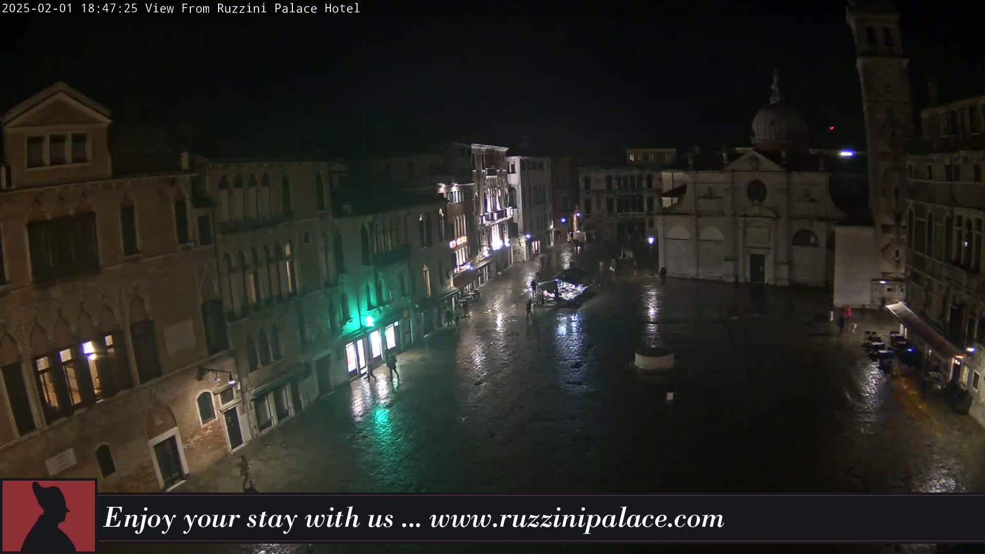 A nighttime, rain-slicked Venetian square is seen from above, showing several buildings, including a church, with a few people visible.