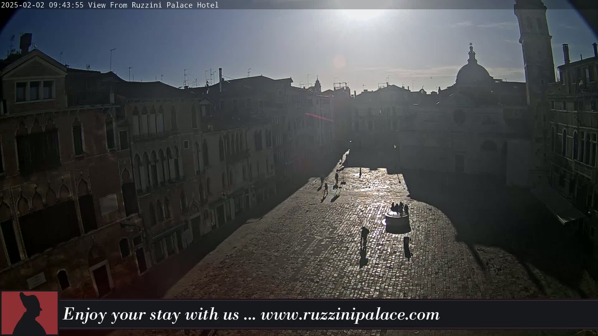 A sunlit cobblestone square in a city, surrounded by old buildings, with a few people visible as silhouettes.