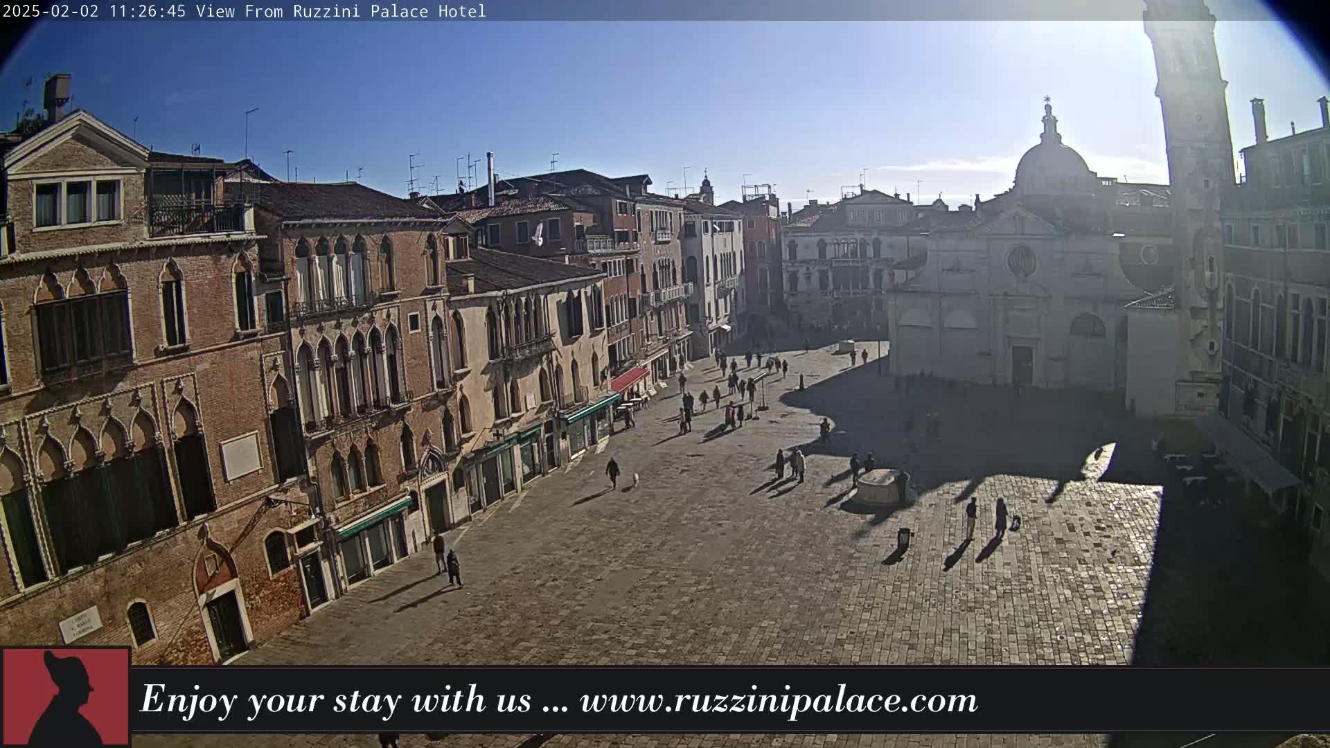 A sunny day reveals a large, mostly empty square in Venice, surrounded by old brick buildings, with a few people walking around and their shadows cast on the ground.