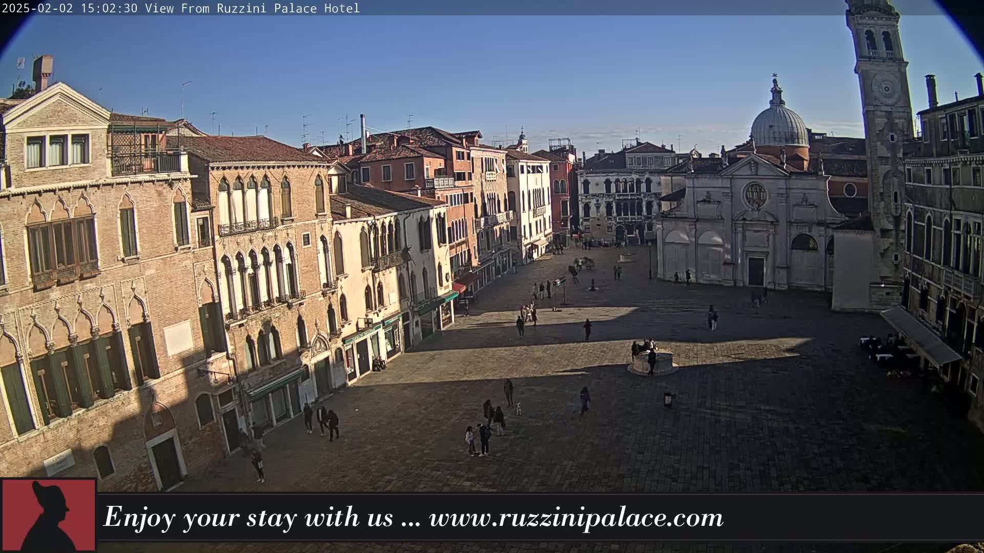 A sunny, daytime view of a large, mostly empty plaza in Venice, Italy, surrounded by historic buildings, including a church and a tall tower.