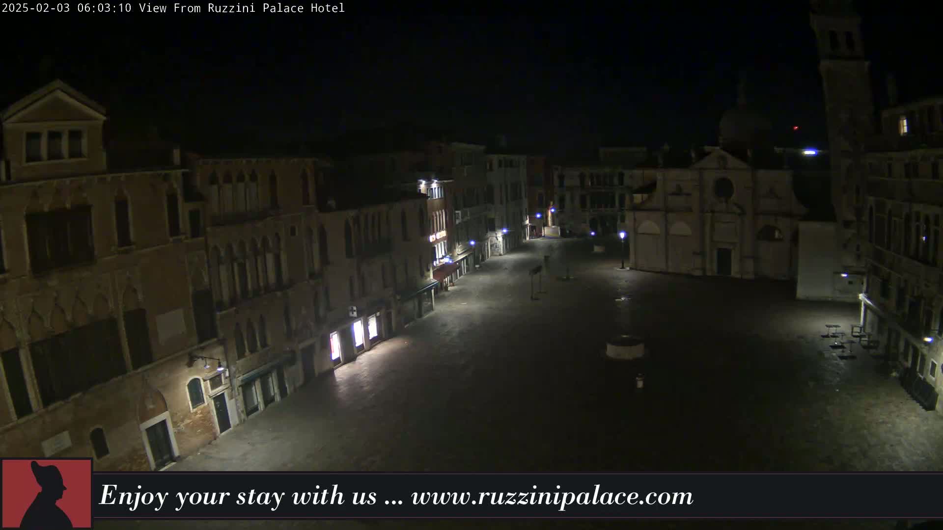 A nighttime, high-angle view of a mostly empty, cobblestone plaza surrounded by dimly lit, historic-looking buildings in Venice.