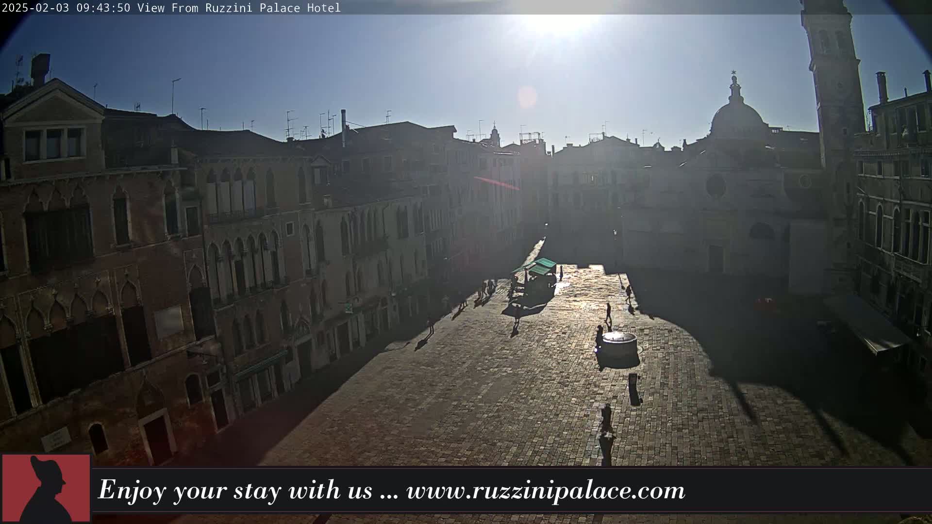 A sunny day reveals a large, mostly empty cobblestone square in Venice, surrounded by tall, historic buildings casting long shadows.