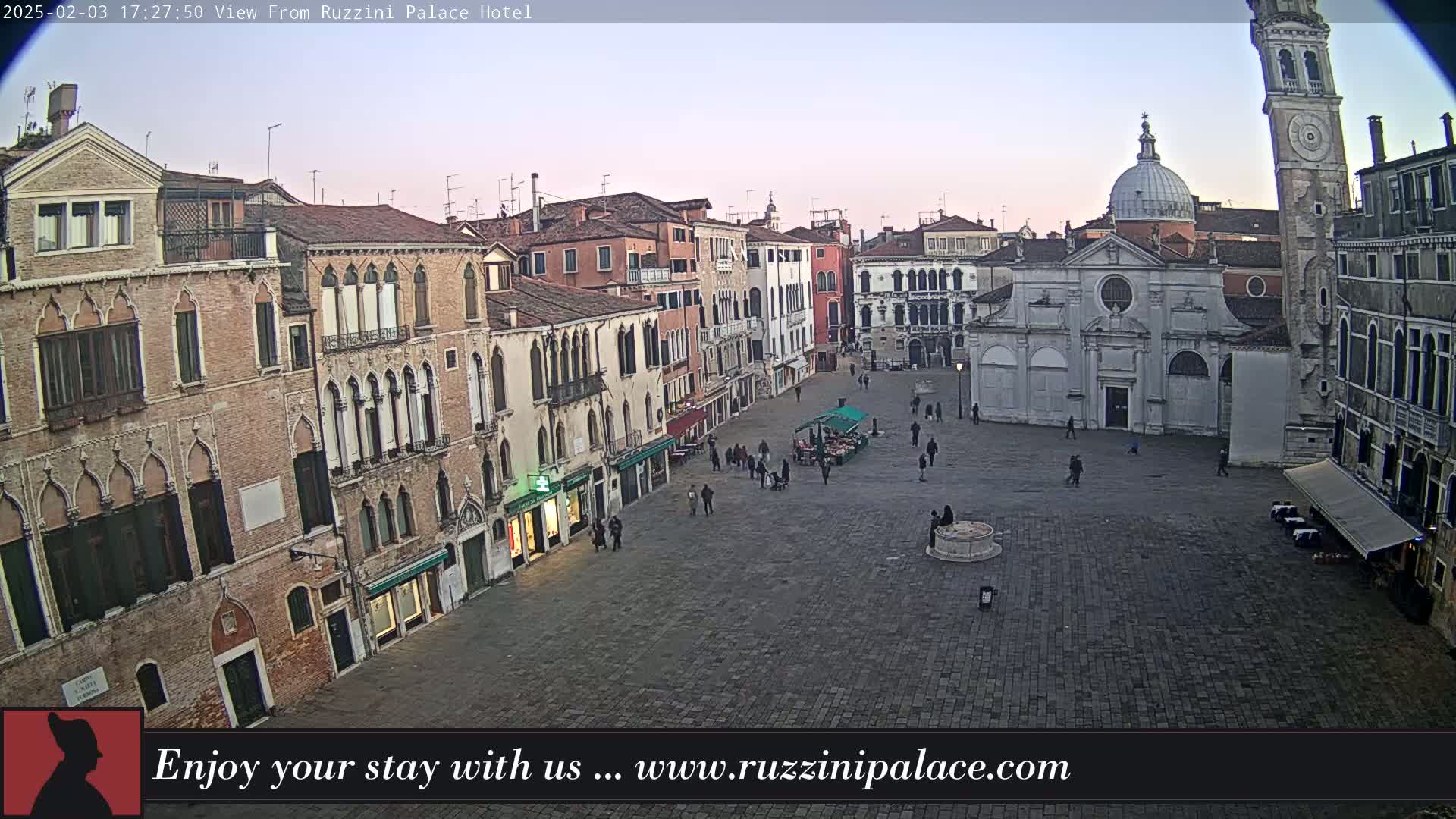 A wide shot of a cobblestone square in Venice, Italy, at dusk, surrounded by old buildings, with a few people sparsely scattered throughout the area.