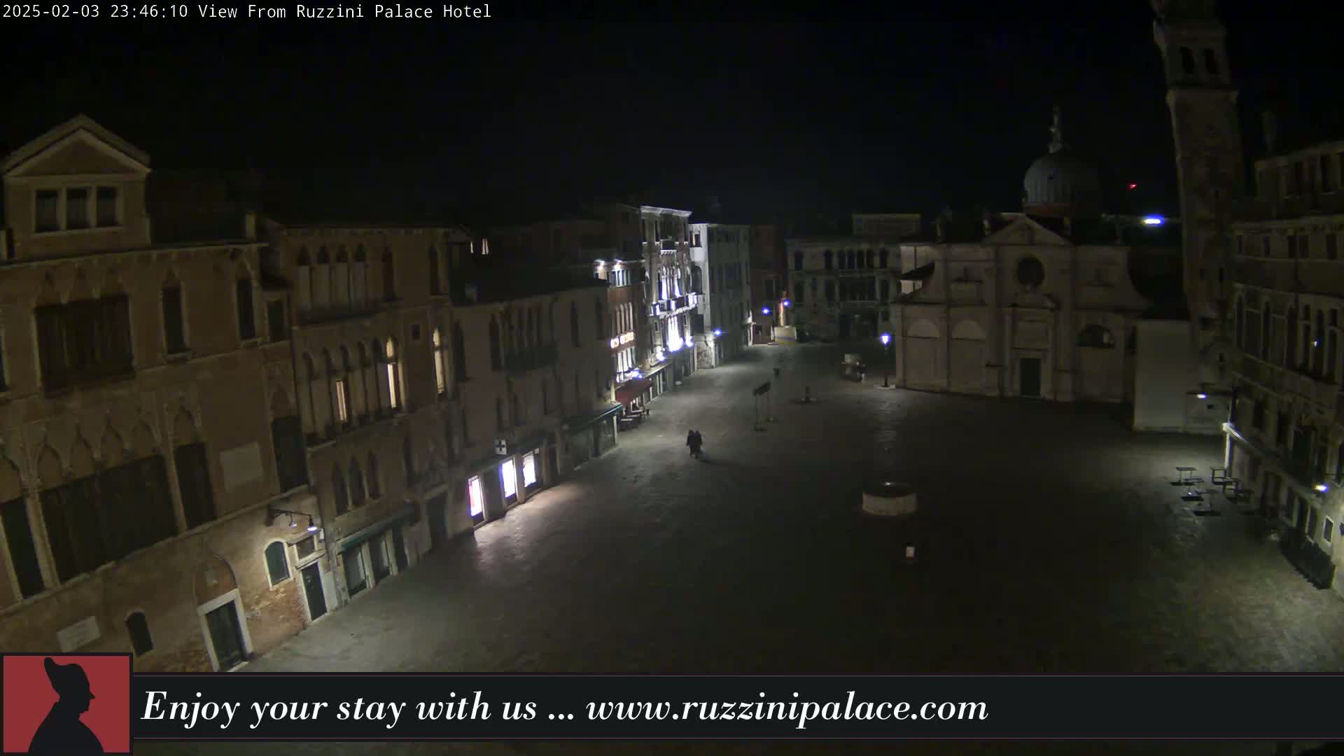 A nighttime, outdoor view of a dimly lit European-style square surrounded by buildings, with a few people visible in the distance.
