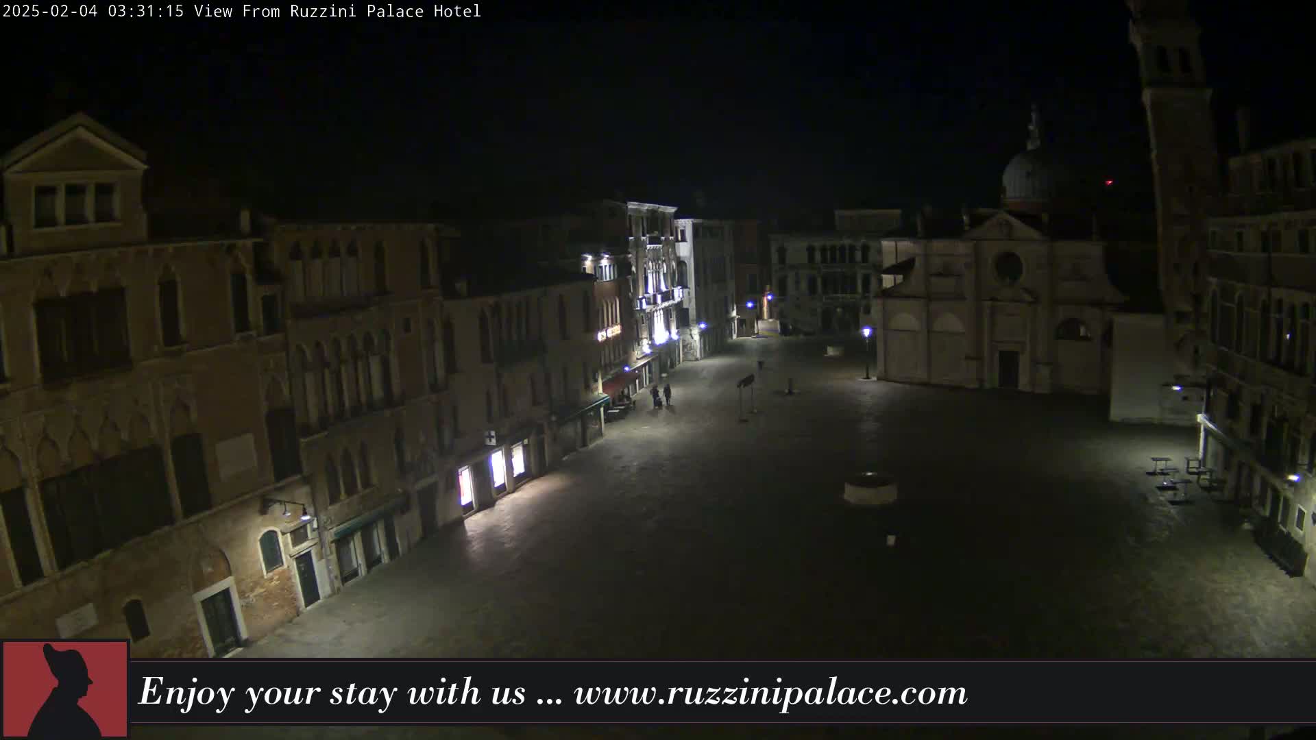 A nighttime, high-angle view of a dimly lit, empty square in Venice, surrounded by historic buildings, with a church visible in the background.