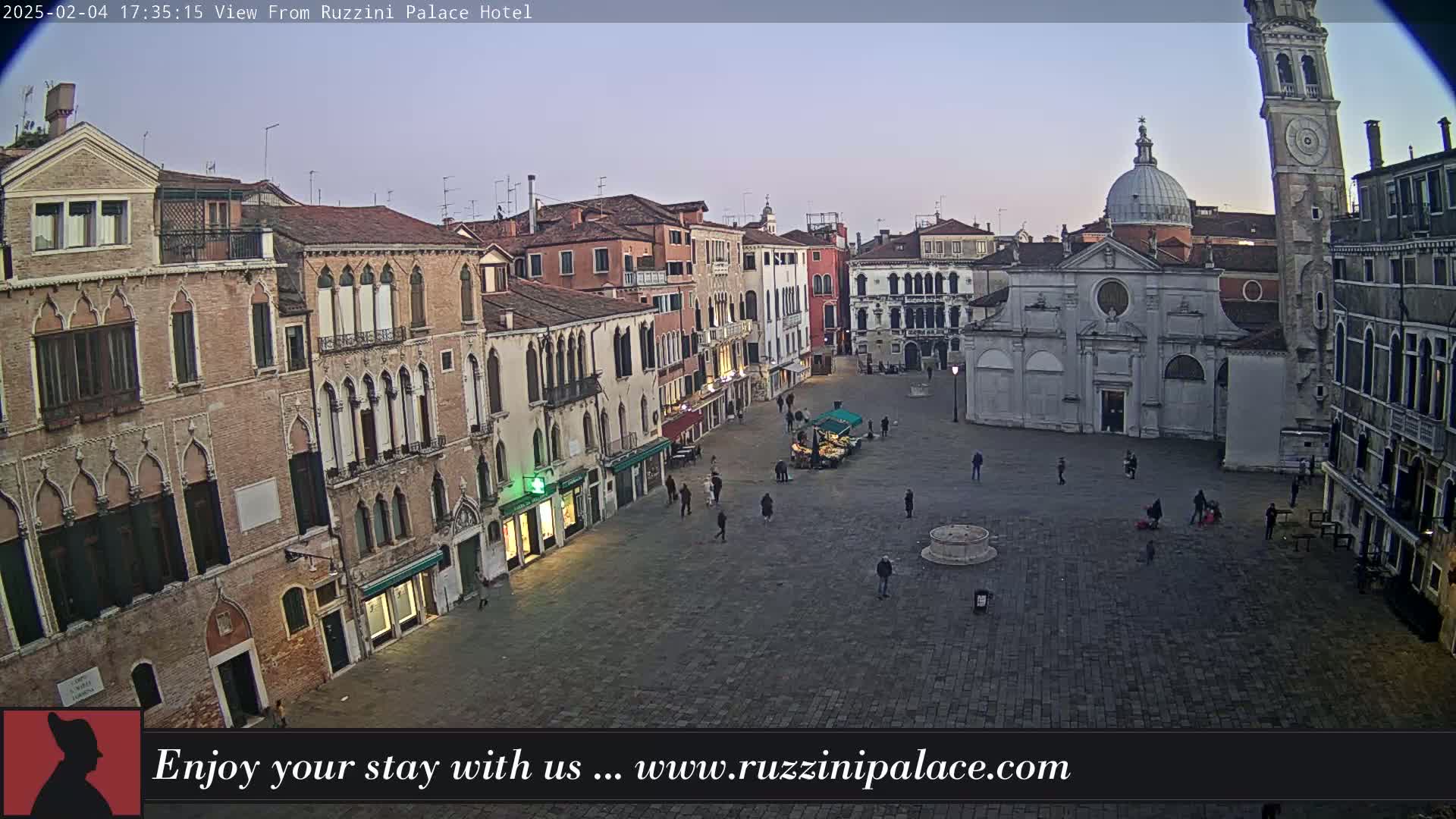 A twilight view shows a large, mostly empty square in Venice, Italy, surrounded by aged buildings, including a church and a tall bell tower.