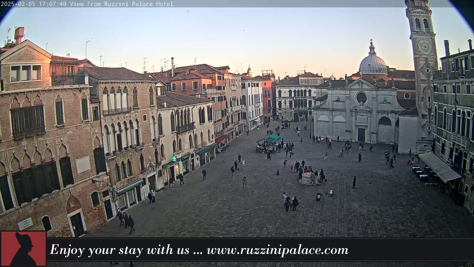A large, paved square in Venice, Italy, is sparsely populated with people, surrounded by historic buildings under a clear, twilight sky.