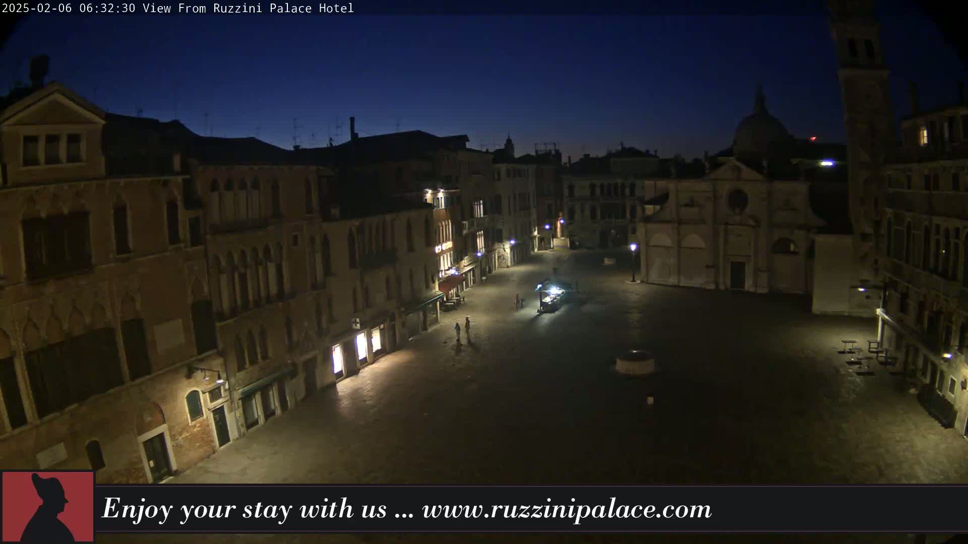 A nighttime, dimly lit view of a large, empty square in Venice, surrounded by old buildings under a dark blue sky.