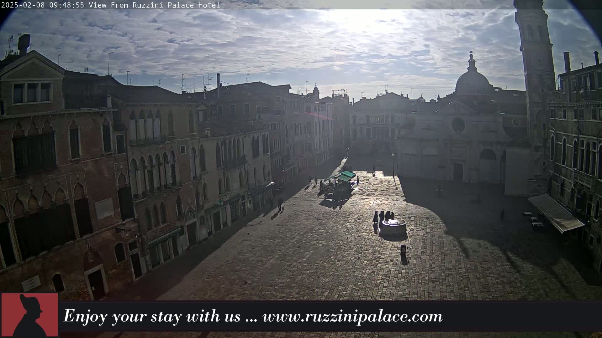 A sunlit, paved square in a city, surrounded by old buildings, under a partly cloudy sky.