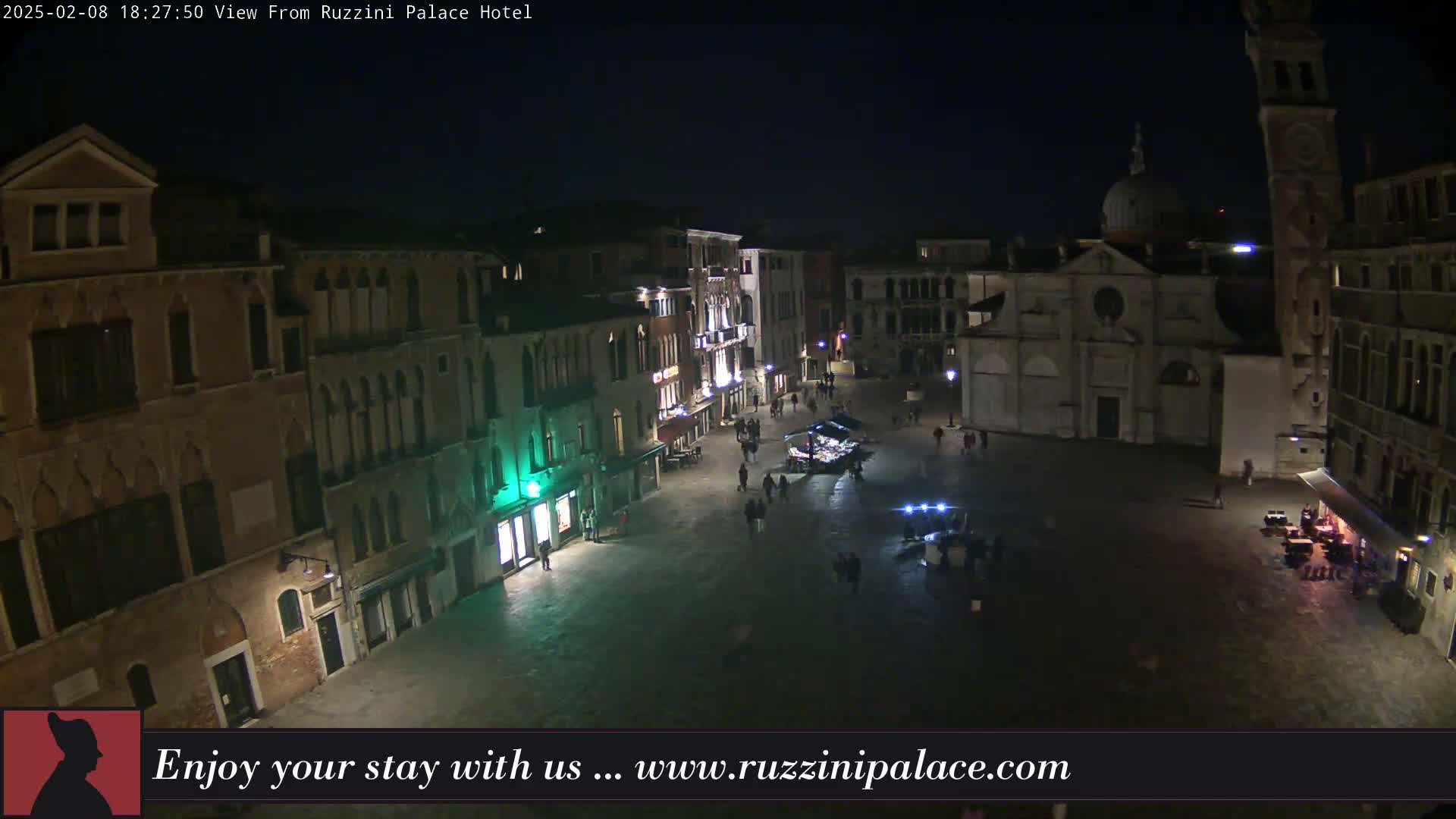 A nighttime view of a dimly lit European-style square with several buildings, a few pedestrians, and a church.