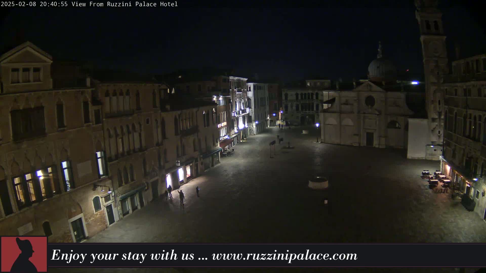 A nighttime, high-angle view of a sparsely populated square in Venice, Italy, surrounded by old buildings, with a church visible in the background.
