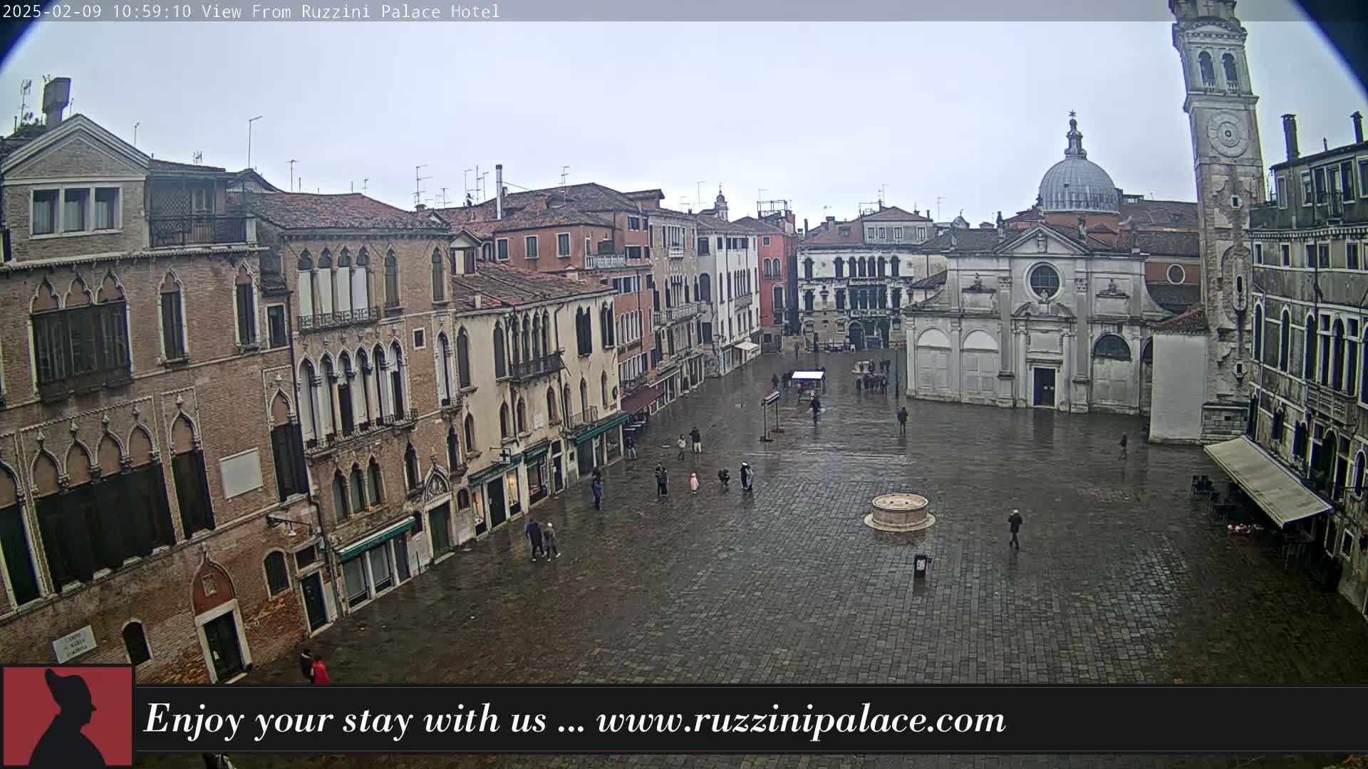A wet, stone plaza in Venice is surrounded by old, multi-storied buildings, with a few people sparsely scattered throughout.