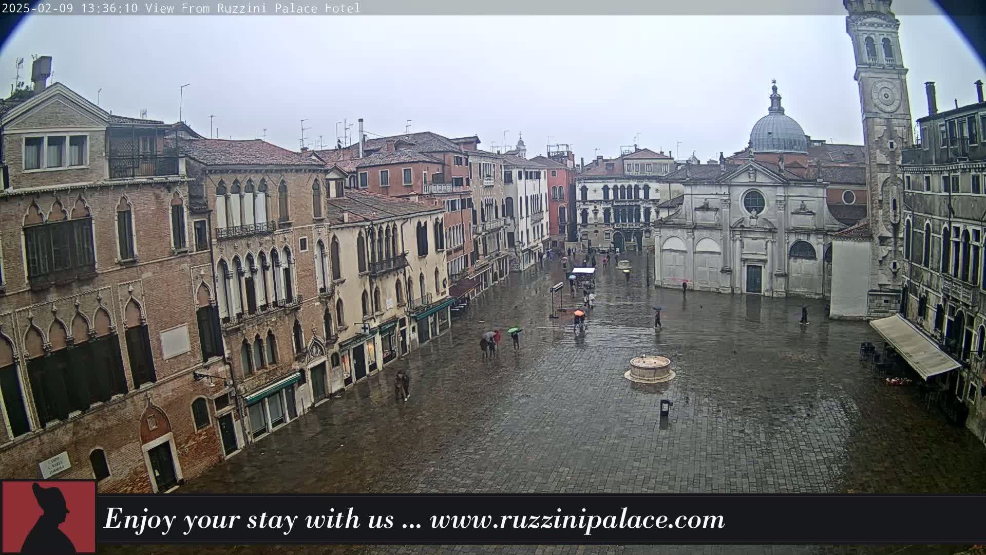 A rain-soaked Venetian piazza, surrounded by historic buildings, is sparsely populated with a few pedestrians carrying umbrellas.