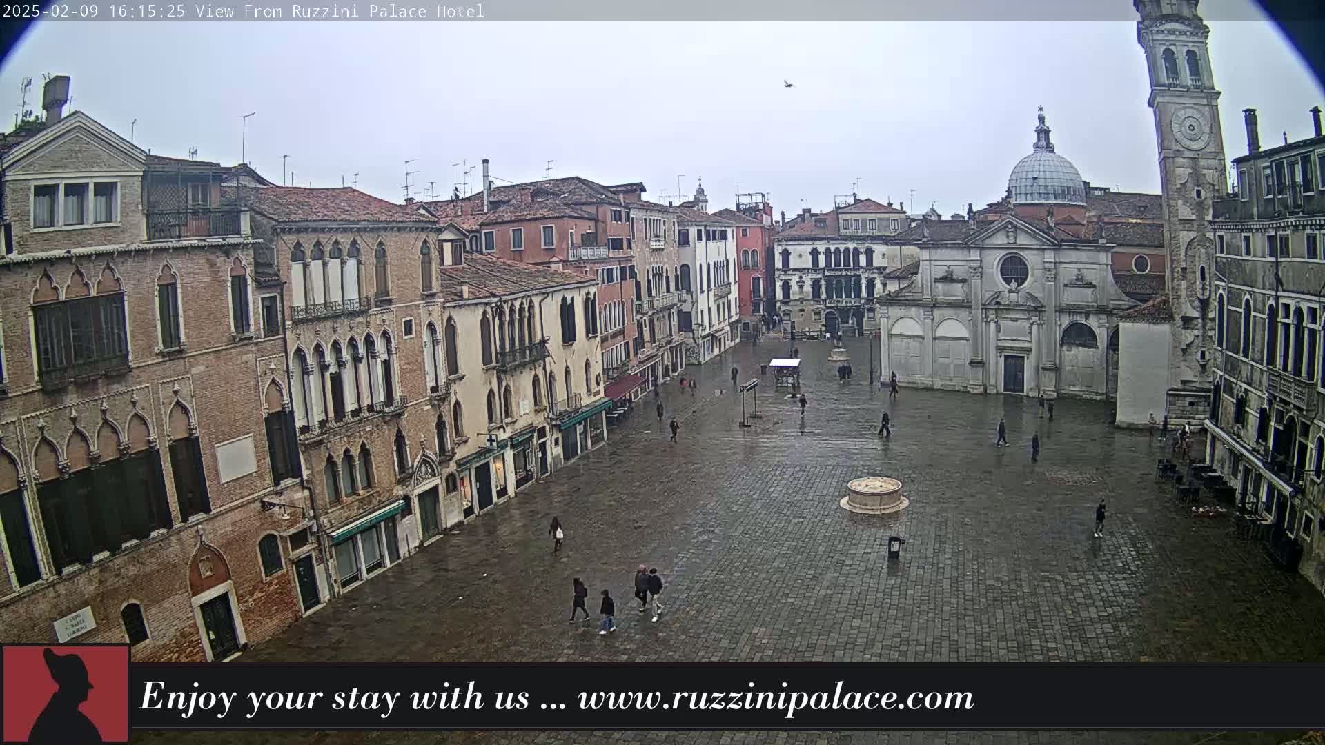 A wet, overcast Venetian square is seen from above, showing several buildings, a church, a clock tower, and a few pedestrians.