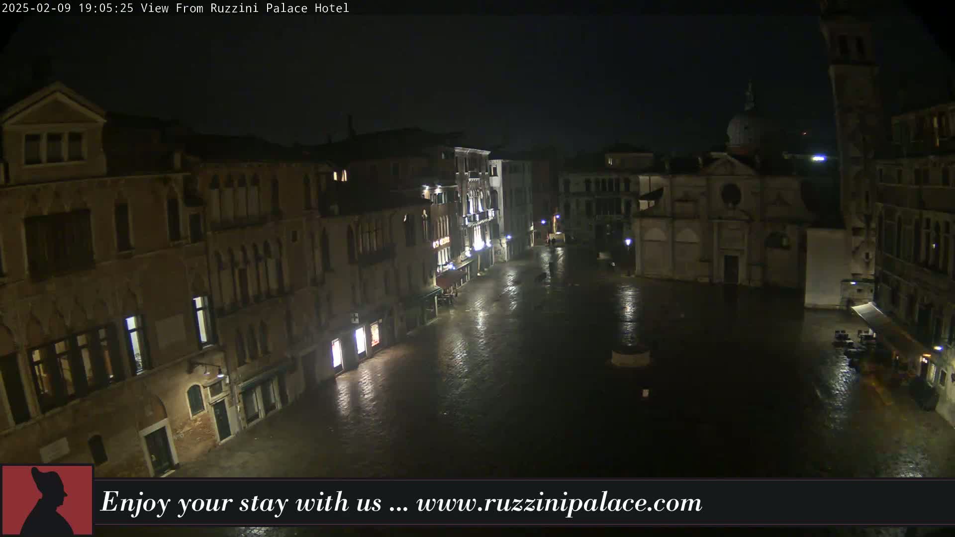 A nighttime view of a rain-soaked Venetian piazza, surrounded by old buildings, one of which is a church with a dome.