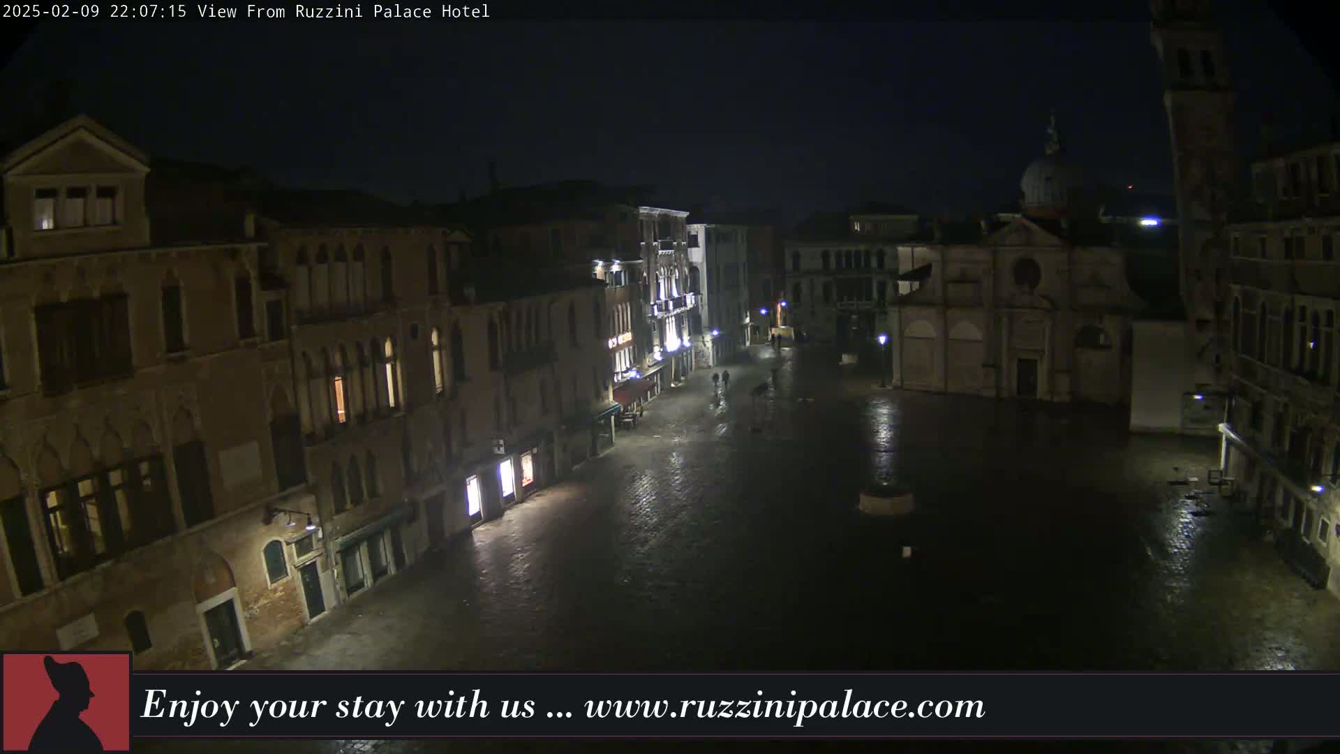 A nighttime view of a wet, empty town square in Venice, Italy, surrounded by old buildings, with a church visible in the background.