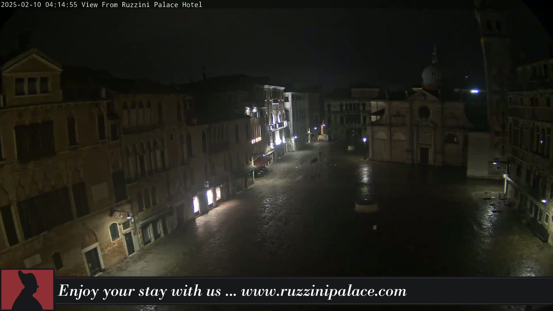 A nighttime view of a mostly empty, cobblestone square in Venice, Italy, under a dark, possibly rainy sky, shows several old buildings lining the square, including a church with a dome.