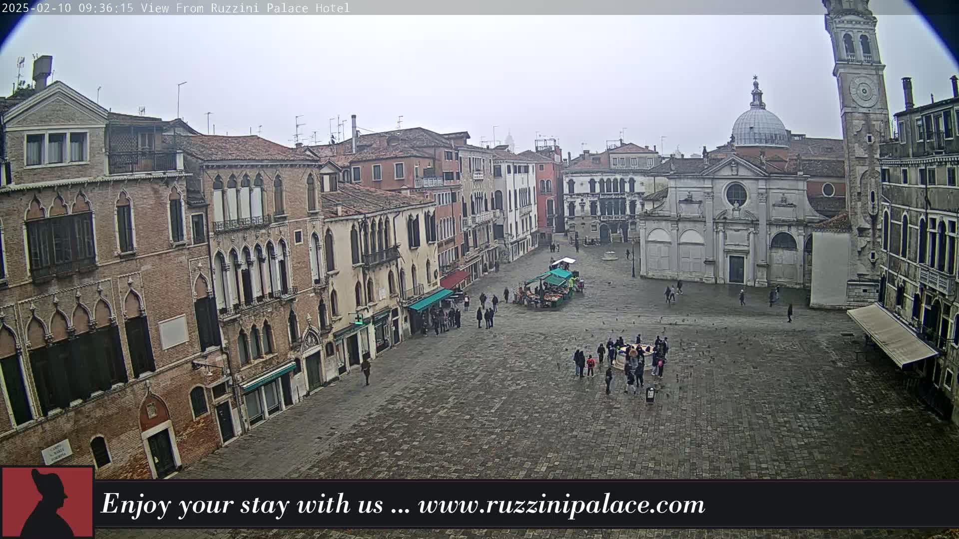 A large, cobblestone square in a city is sparsely populated with people, surrounded by old, multi-story buildings under an overcast sky.