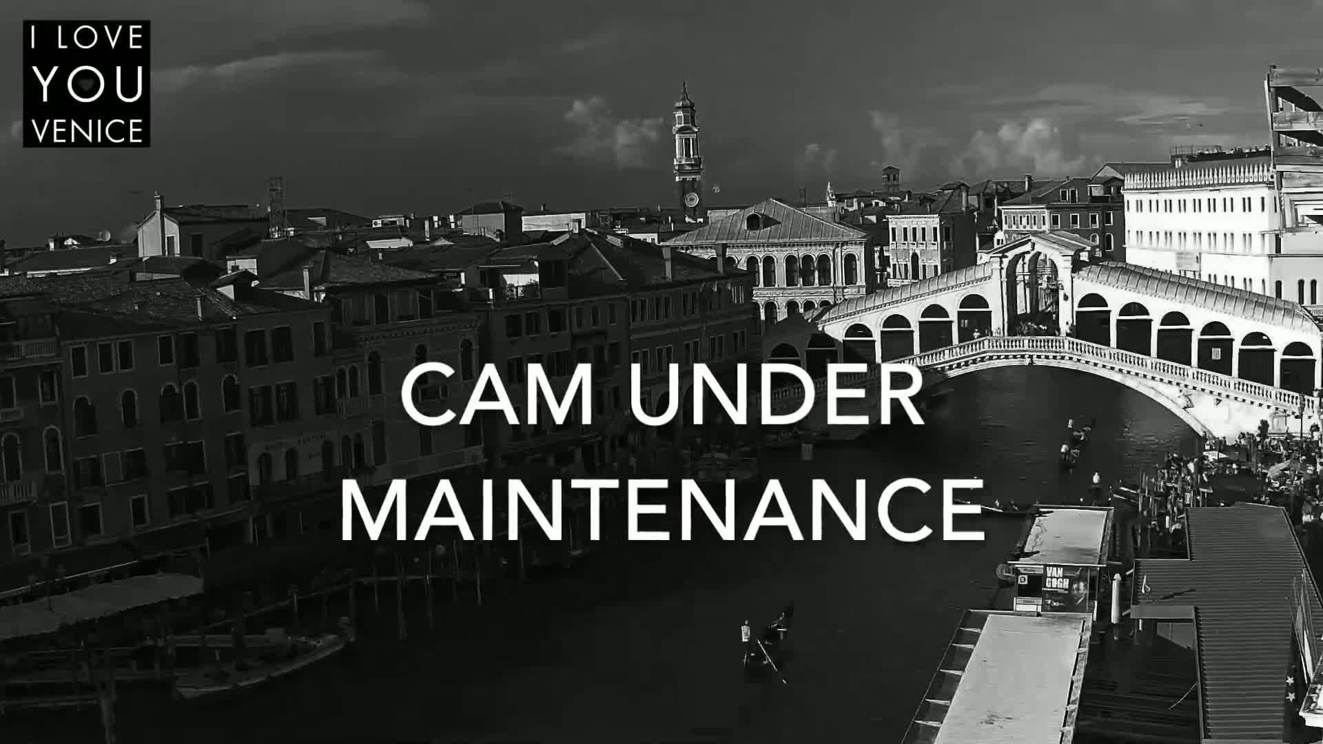 A black and white, high-angle view shows a bridge spanning a canal in Venice, with buildings lining the waterway under a partly cloudy sky.