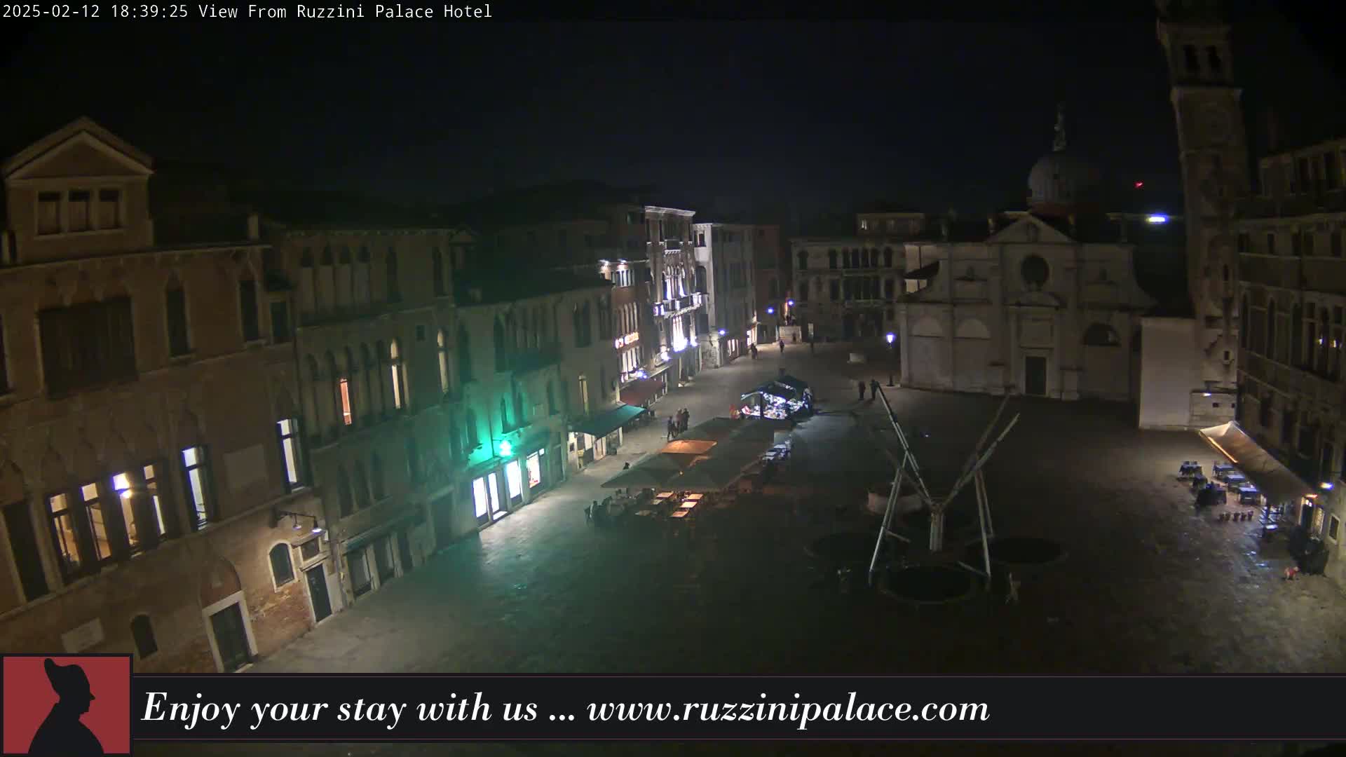 A nighttime, aerial view of a dimly lit Venetian piazza, featuring several buildings, a church, and sparse outdoor seating.