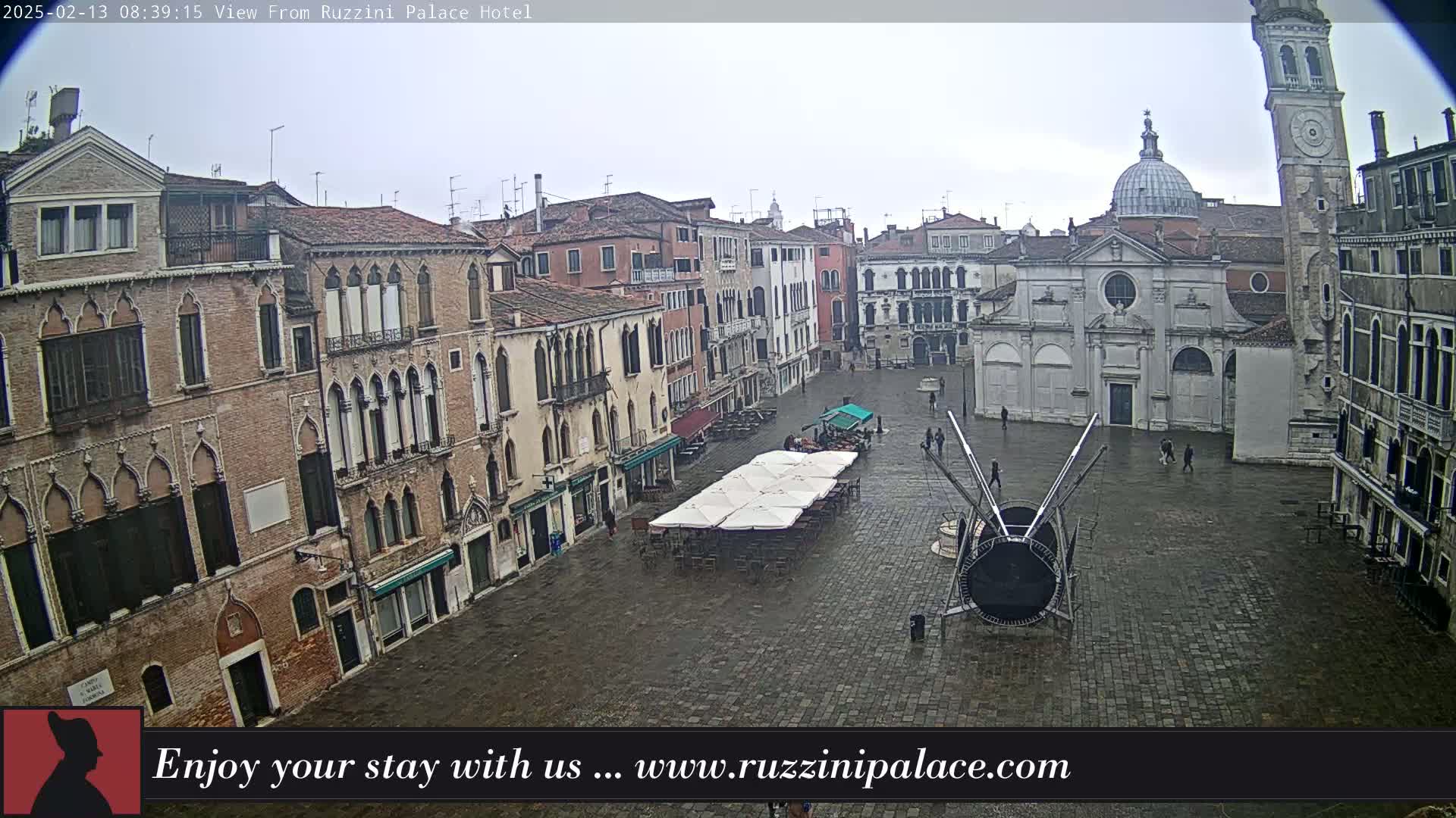 A wet, overcast Venetian square is shown, featuring several historic buildings, an outdoor café with umbrellas, and a large modern art installation.
