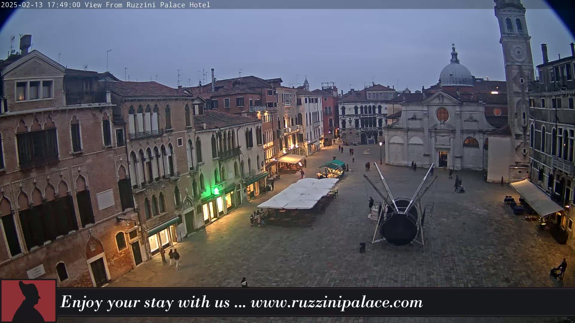 A twilight view of a large, paved square in Venice, Italy, surrounded by historic buildings, with a few people and market stalls visible.