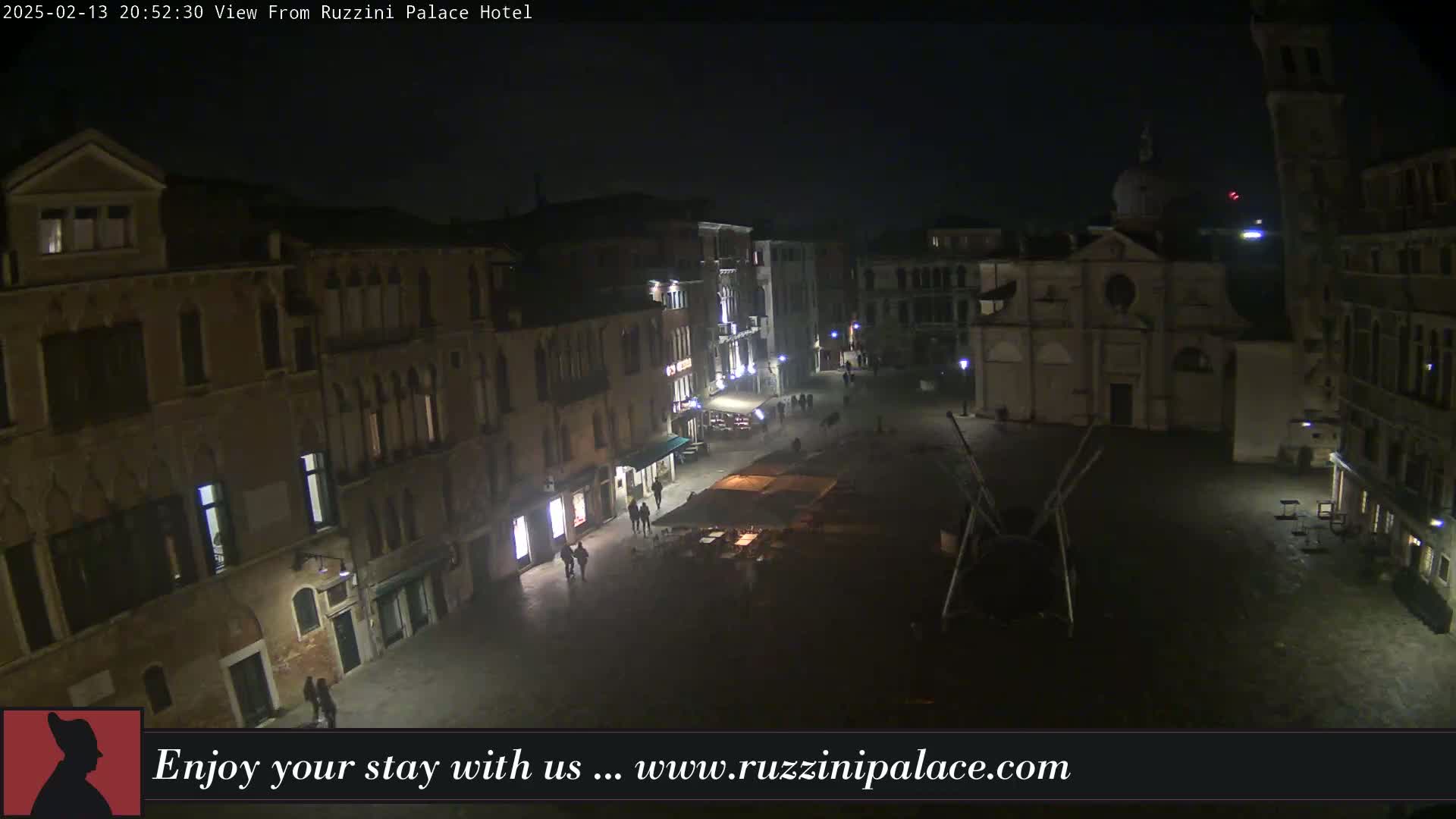 A nighttime, high-angle view shows a sparsely populated town square in Venice, Italy, with buildings lining its perimeter.