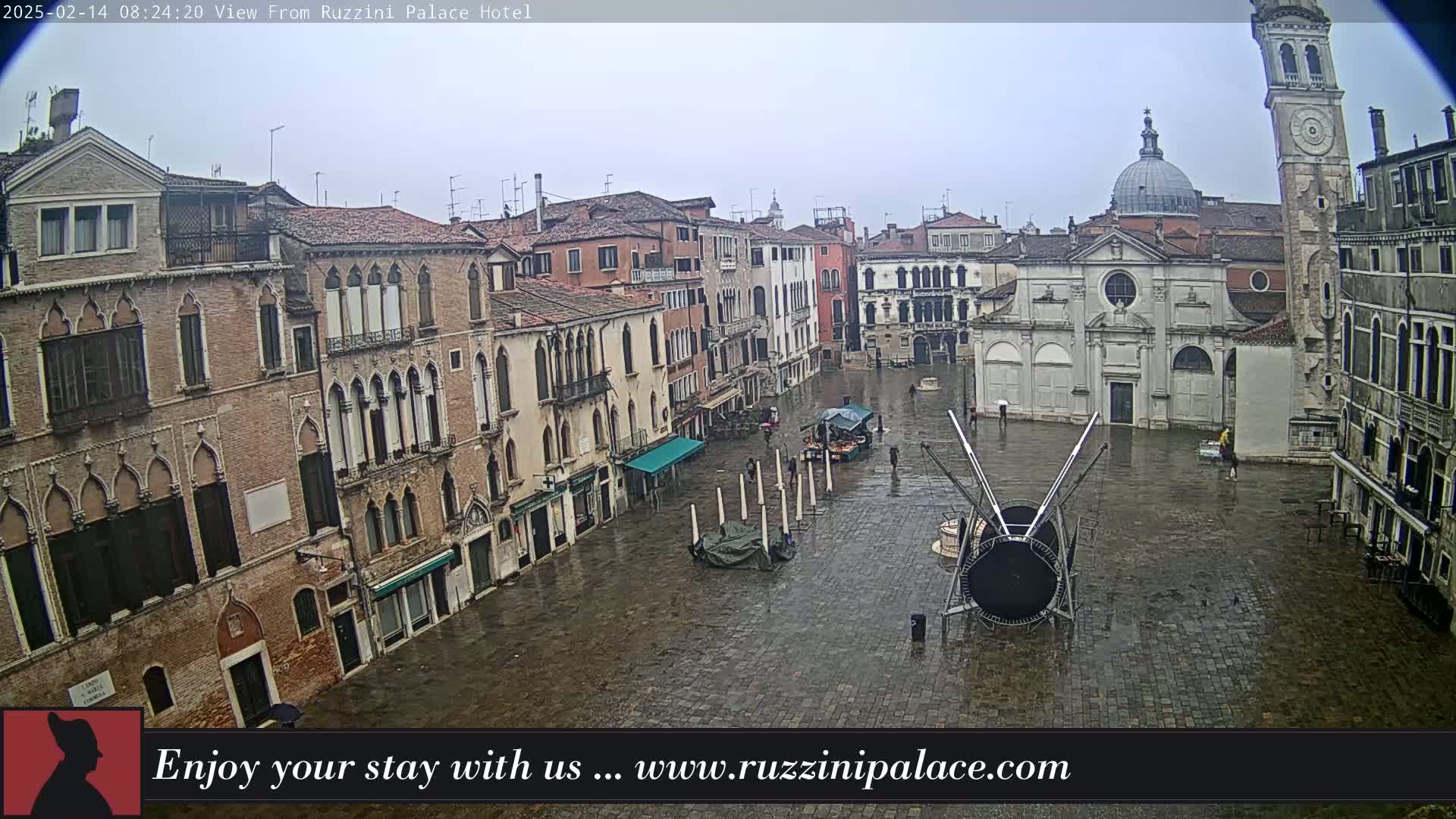 A wet, stone plaza in Venice, Italy, is surrounded by old, multi-story buildings, with a church and clock tower visible in the background.