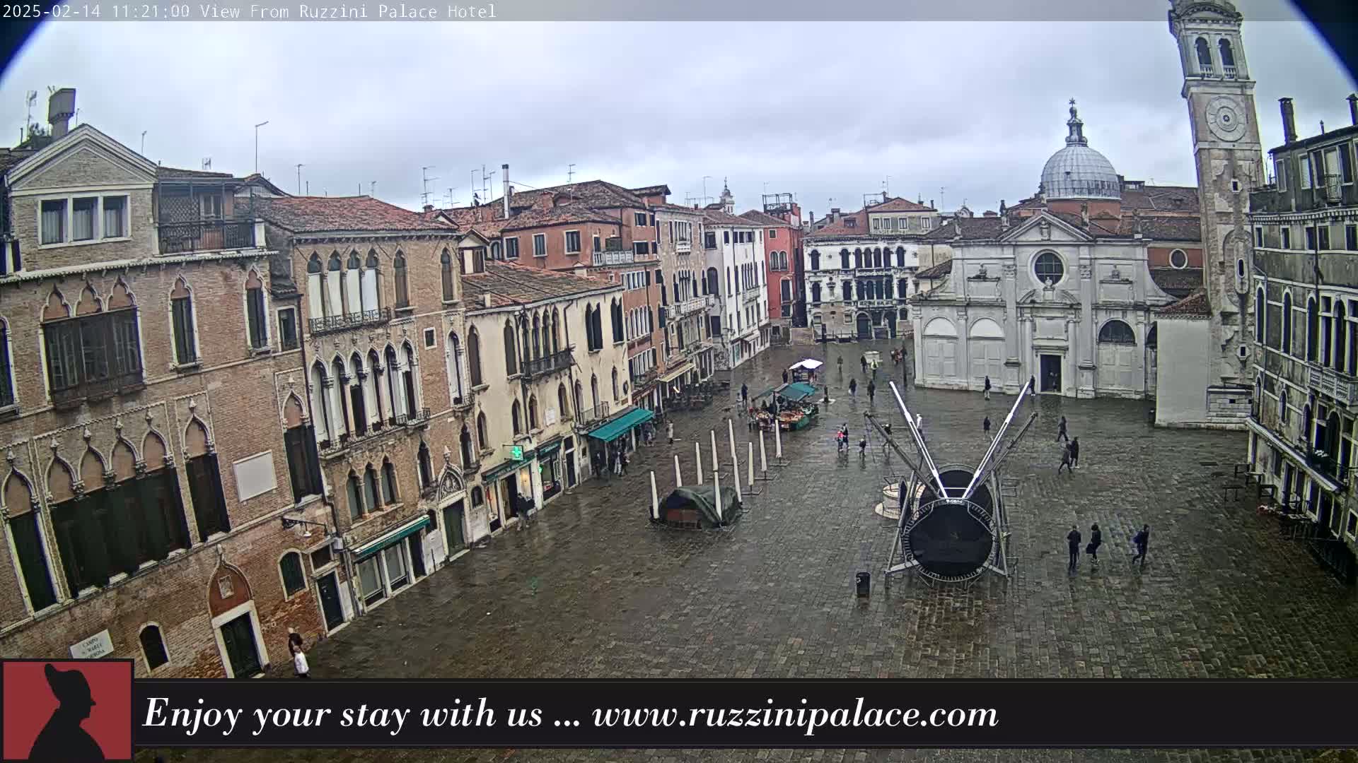 A wet, overcast Venetian square is viewed from above, showing several buildings, a church, and a few pedestrians.