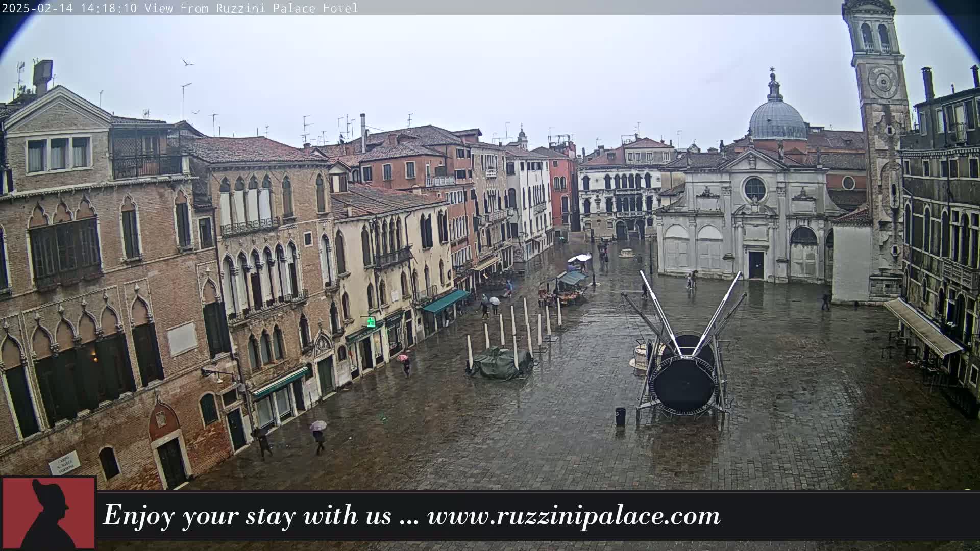 A wet, paved Venetian square is surrounded by old brick buildings, including a church and a tall clock tower, under an overcast sky.
