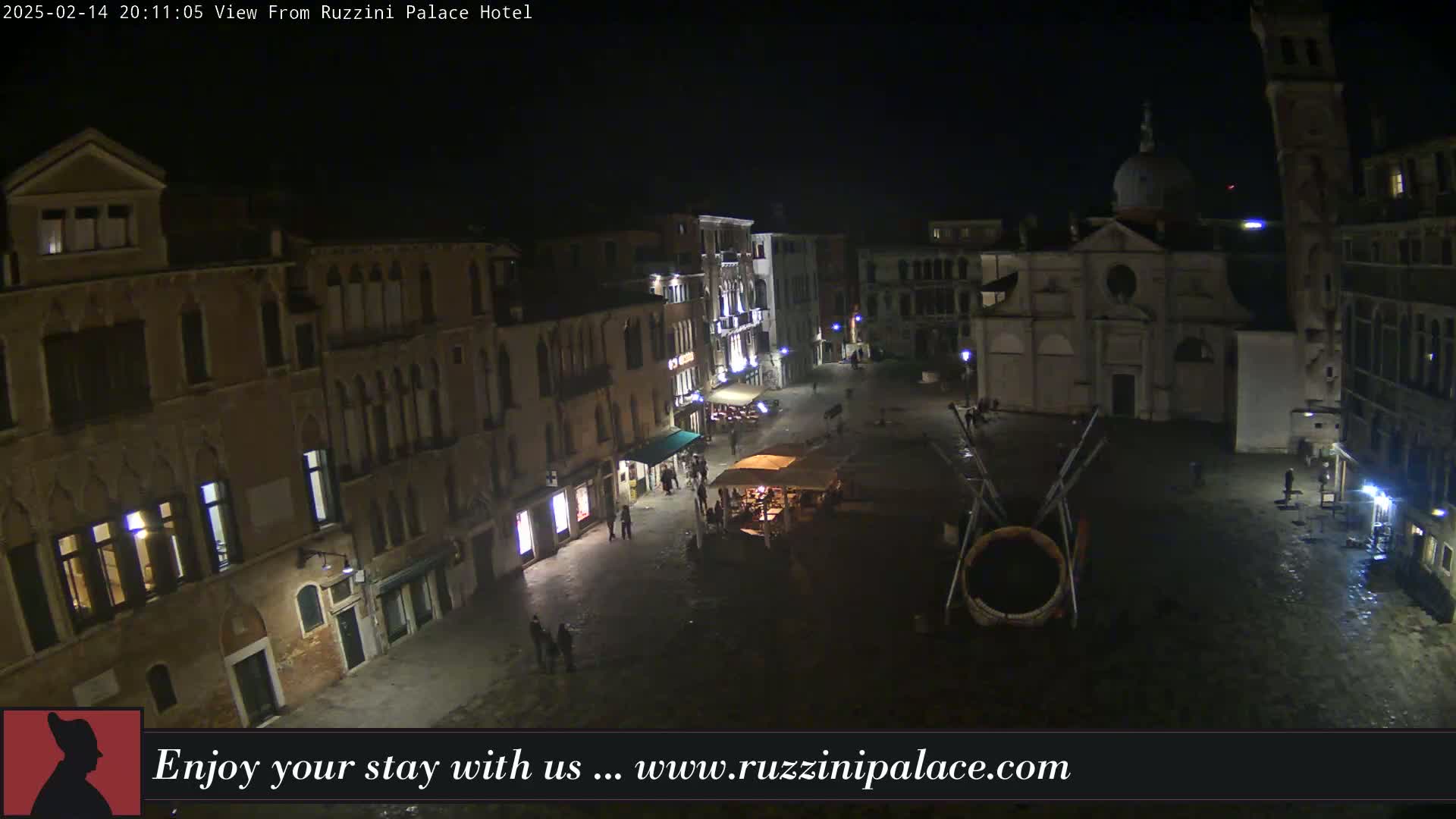 A nighttime, aerial view of a dimly lit, cobblestone square in Venice, featuring several buildings, a church, a few people, and an art installation.