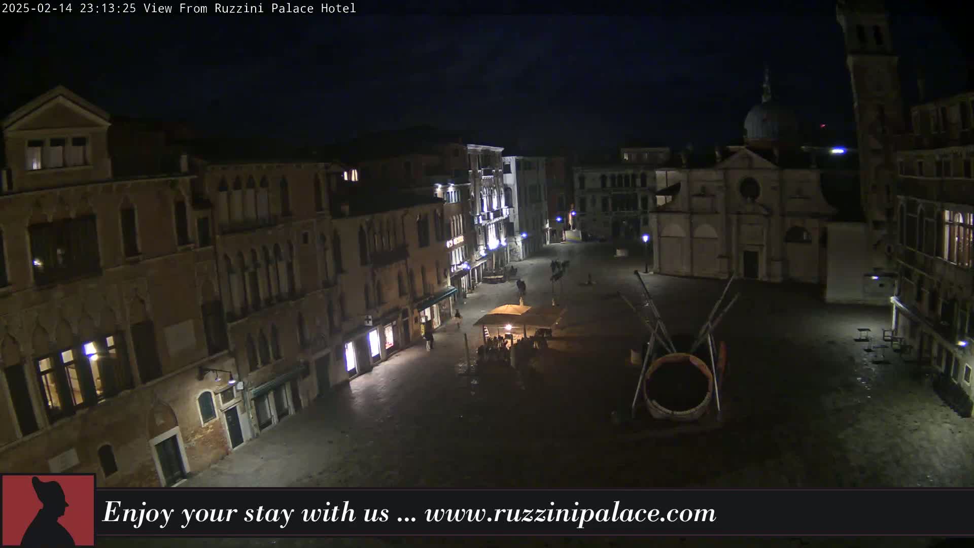 A nighttime view of a dimly lit Venetian piazza, featuring several buildings, a church, and a few people gathered around an outdoor structure.