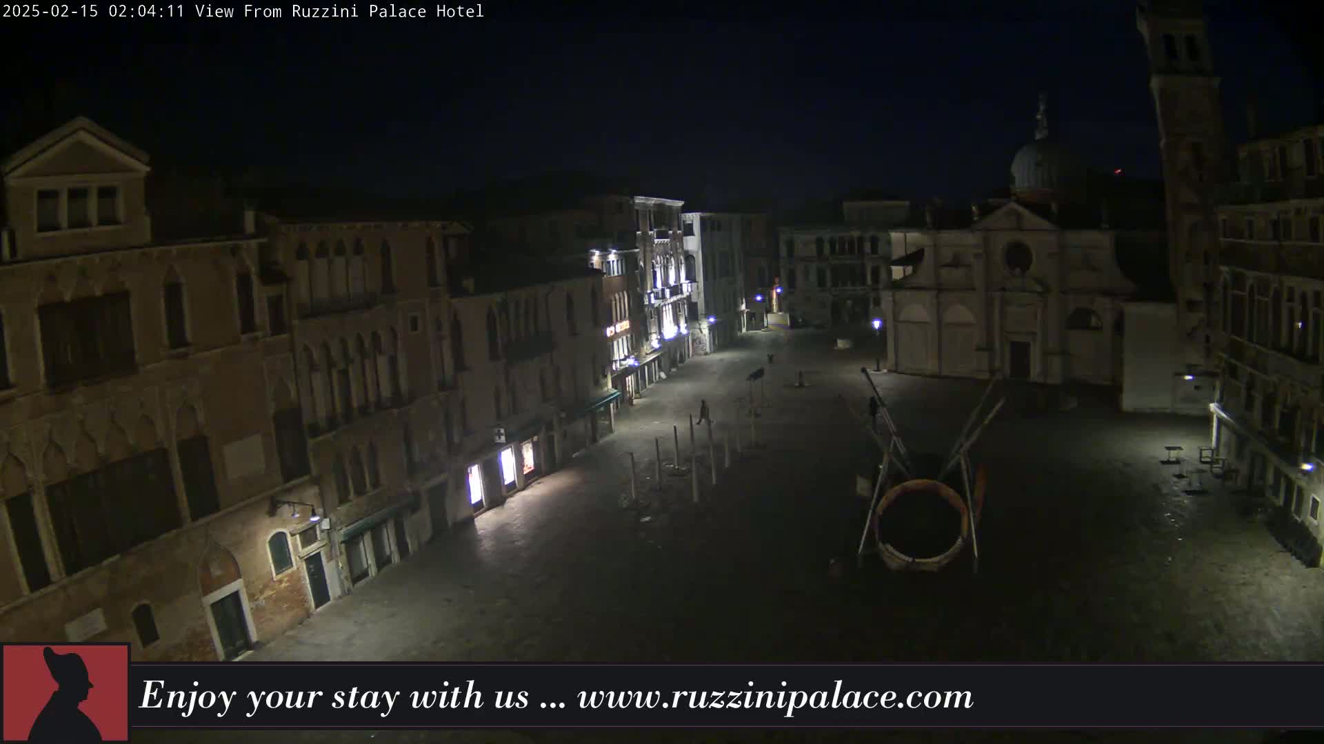 A nighttime, deserted Venetian square is illuminated by streetlights, featuring several buildings, a church, and a large circular sculpture.