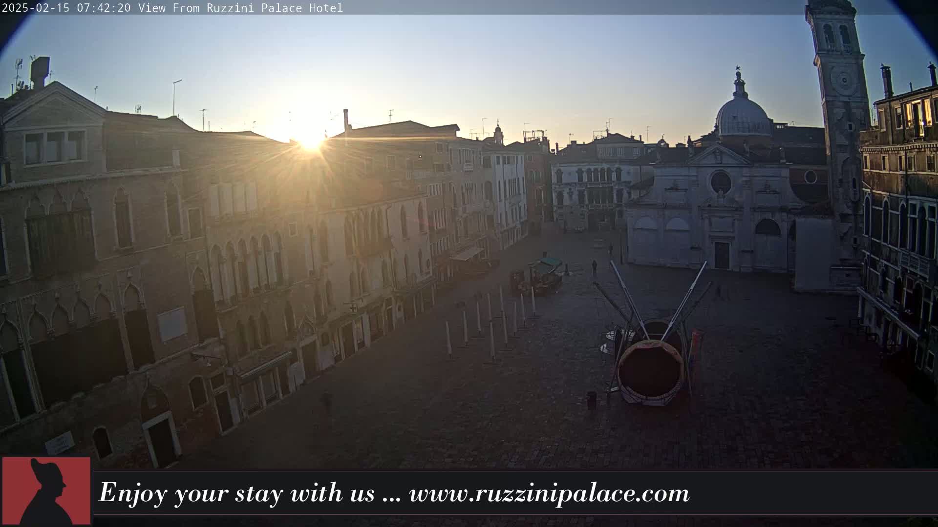 A sunlit view of a cobblestone Venetian piazza surrounded by aged buildings, with a church and tall bell tower in the background under a clear sunrise sky.
