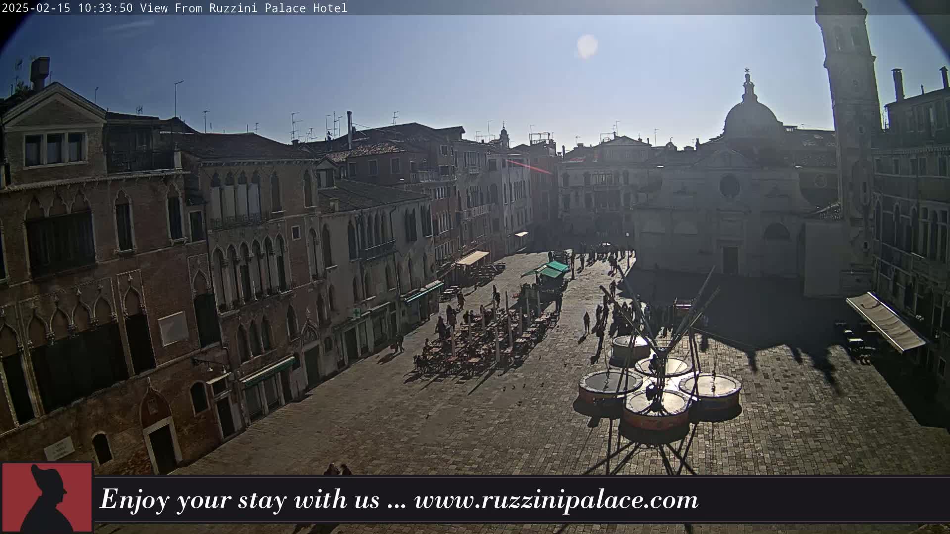 A sunny, high-angle view of a cobblestone plaza in Venice, Italy, shows several buildings surrounding the square with people and outdoor seating visible.