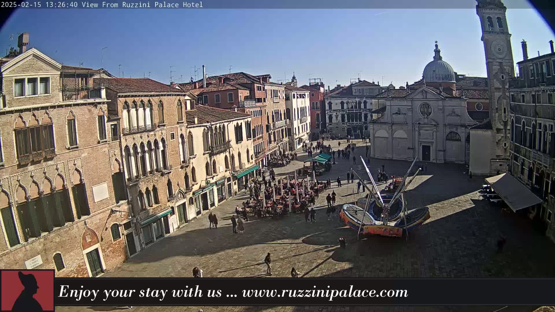 A sunny day reveals a large, paved square in Venice, Italy, filled with people, outdoor seating at cafes, and a small amusement ride, surrounded by historic buildings.