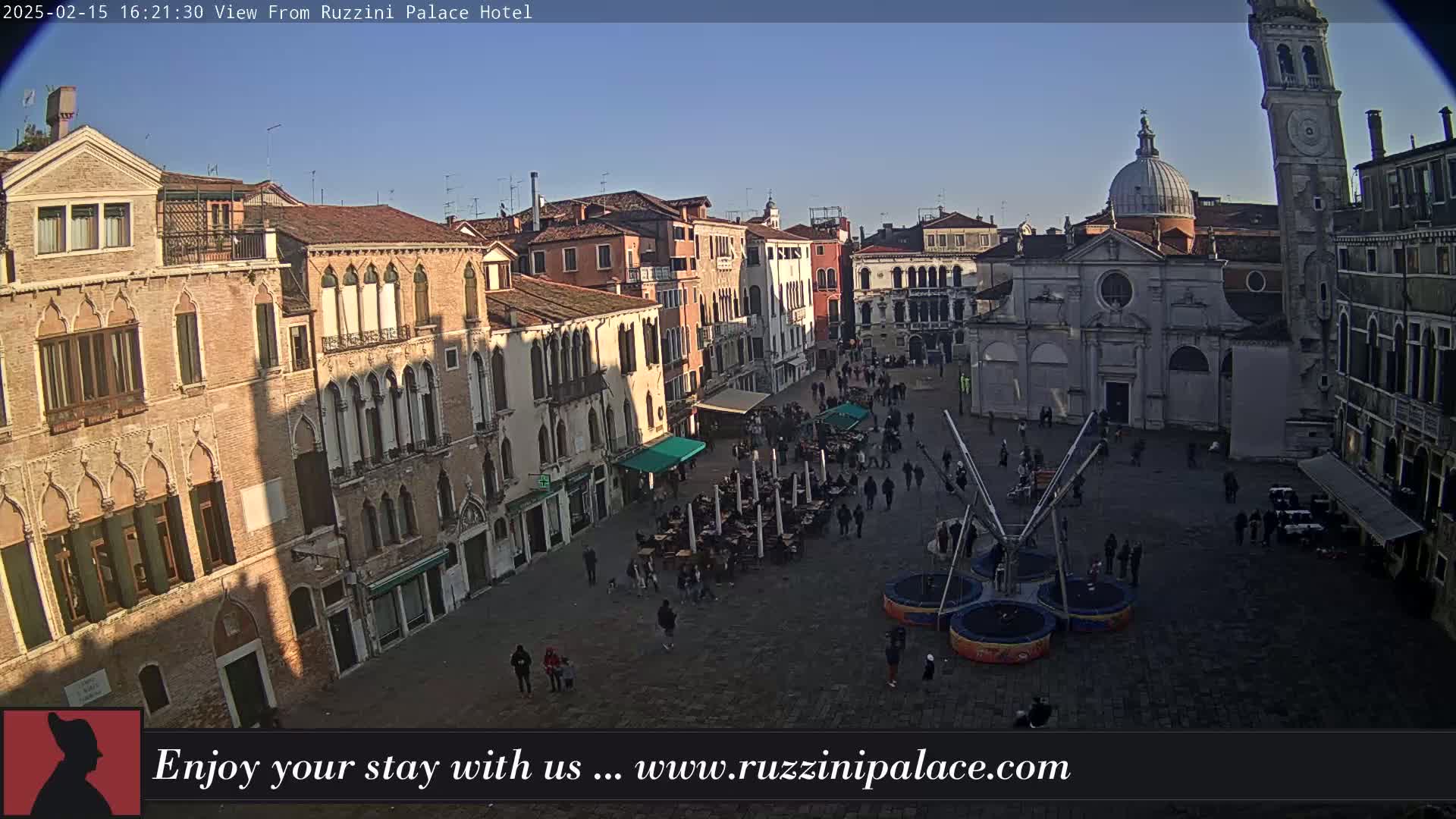 A sunny view of a large plaza in Venice, Italy, filled with people, outdoor seating, and a small trampoline-like structure, surrounded by historic buildings.