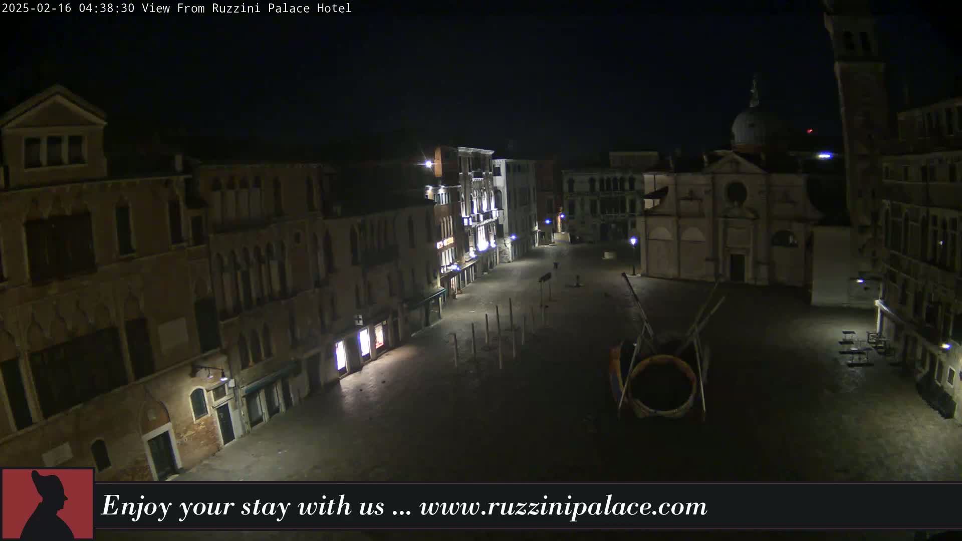 A nighttime view of a deserted, cobblestone square in Venice, Italy, surrounded by old buildings, with a church and a modern art installation visible.