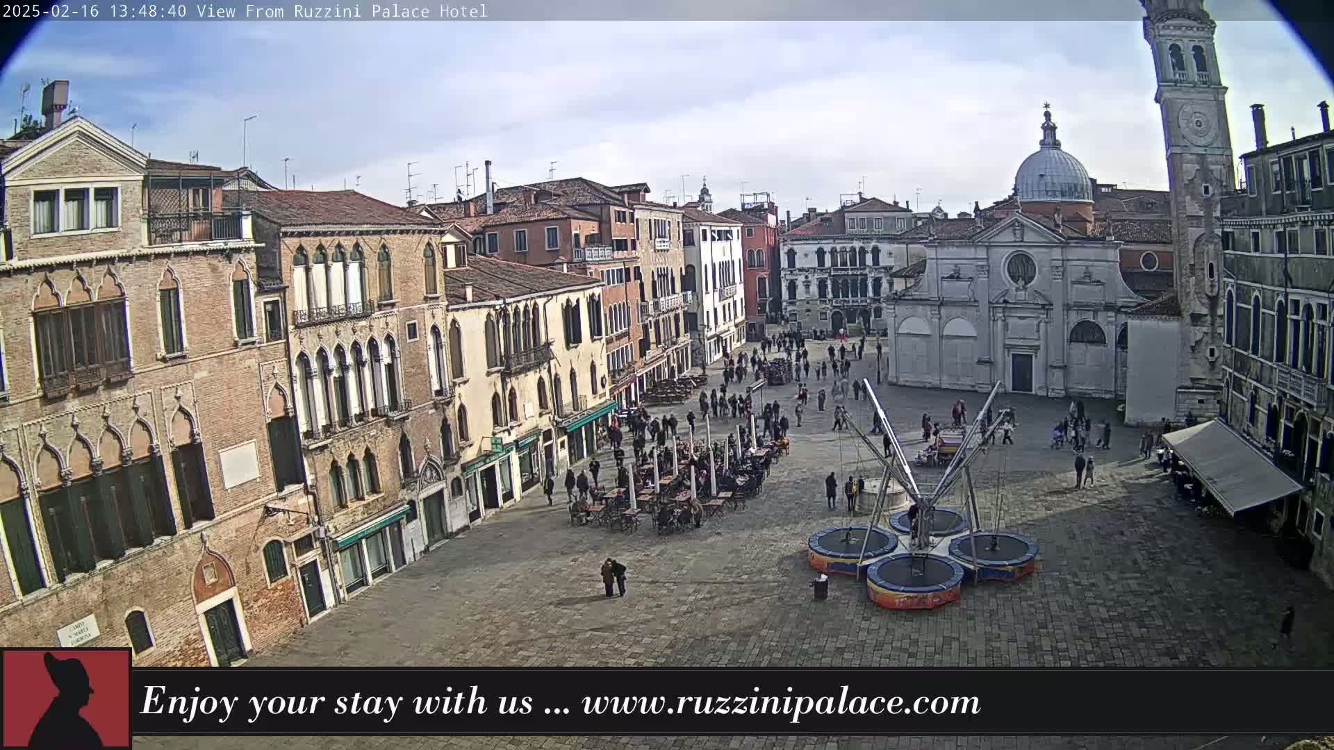 A mostly sunny day reveals an Italian plaza filled with people and buildings, including a church with a dome and a leaning tower, with several trampolines set up in the square.